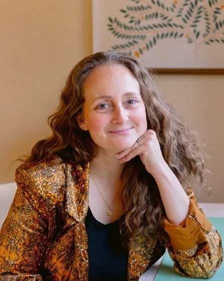 A woman with curly hair, smiling softly, resting her chin on her hand, sitting at a desk with a piece of paper and a pen in front of her, in a room with a yellow wall and a framed abstract art piece.
