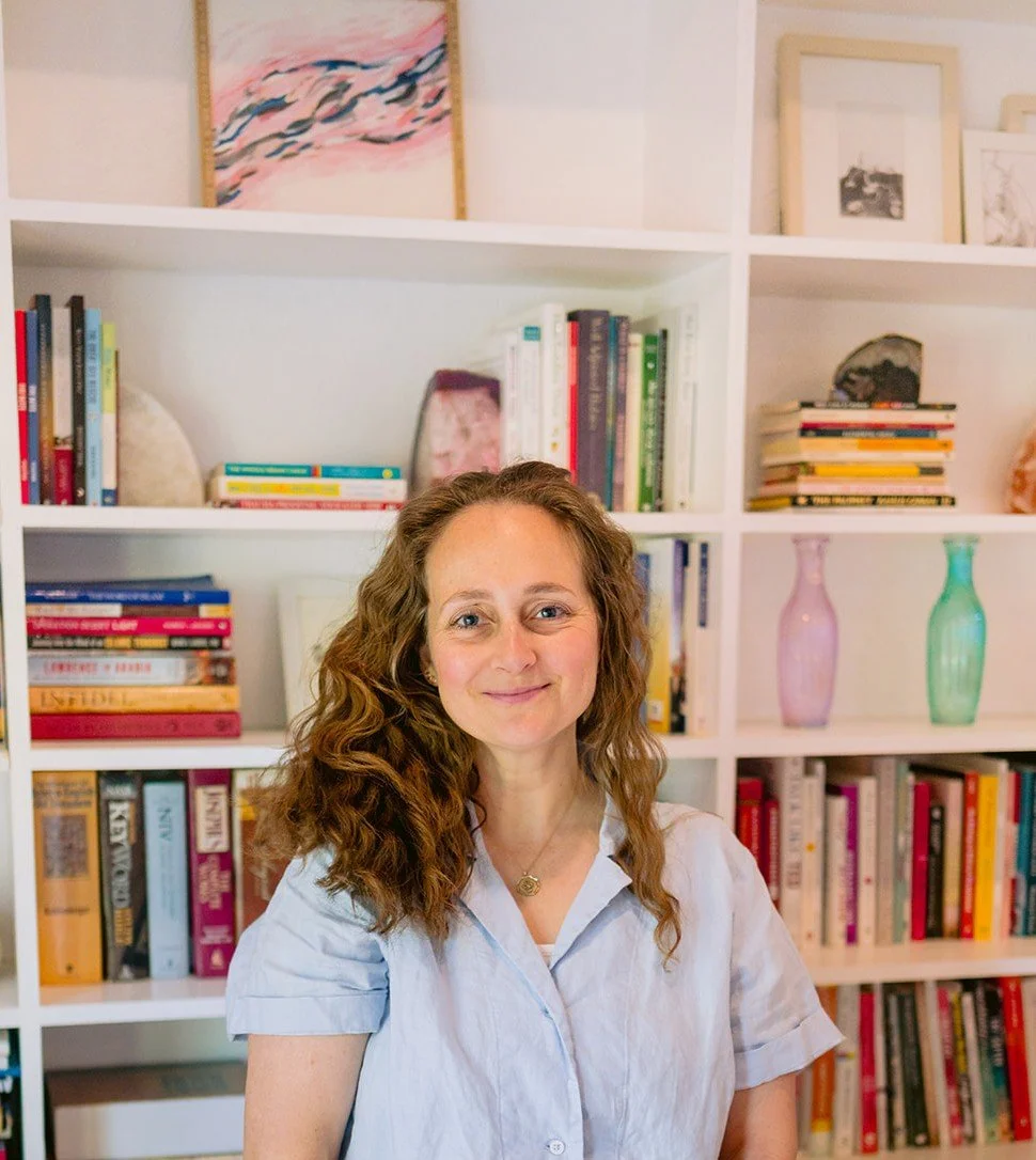 A woman with curly dark blonde hair smiling in front of a white bookshelf filled with books, vases, and framed pictures.