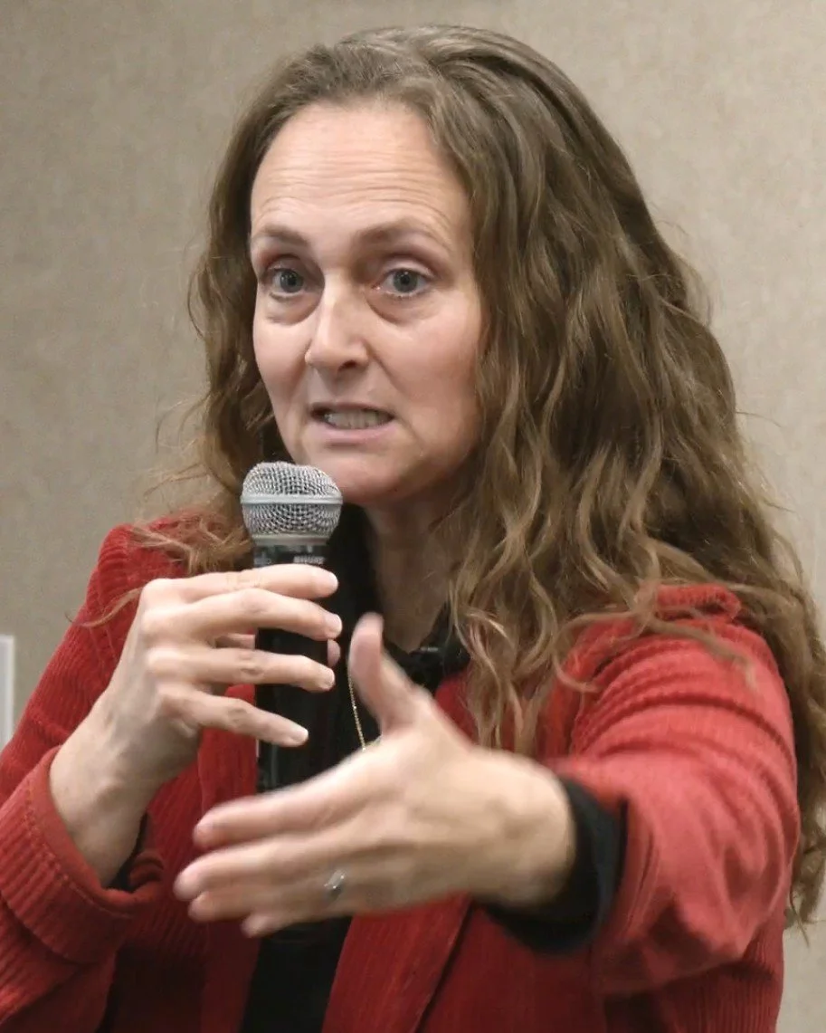 A woman with long, curly hair wearing a red jacket, speaking into a microphone, making a hand gesture.