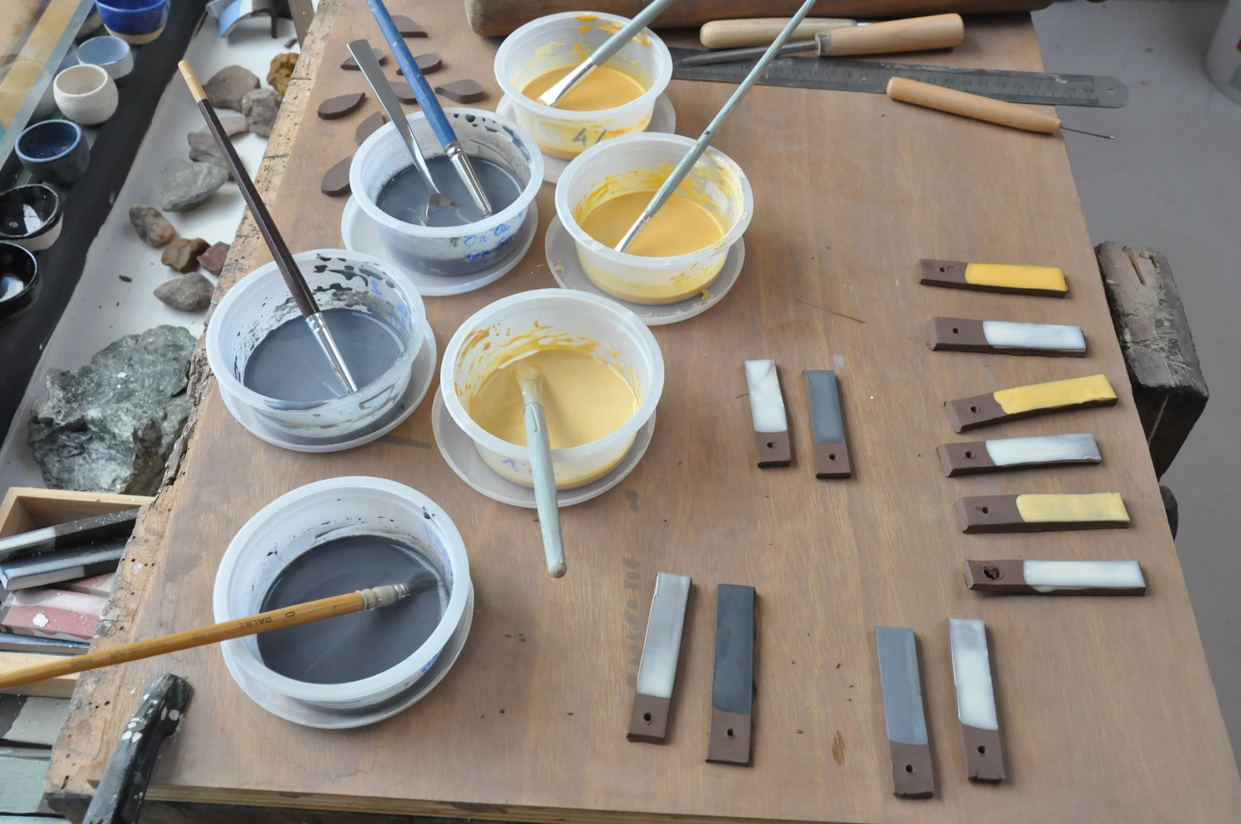 Table de travail d'atelier avec des pots de peintures ou d'émaux en divers coloris, des pinceaux, un couteau à palette, et divers outils pour la sculpture ou la peinture. Sur le côté, des morceaux de céramique ou de bois avec de la peinture appliquée. En arrière-plan, des petits bols et des pierres sur une étagère.
