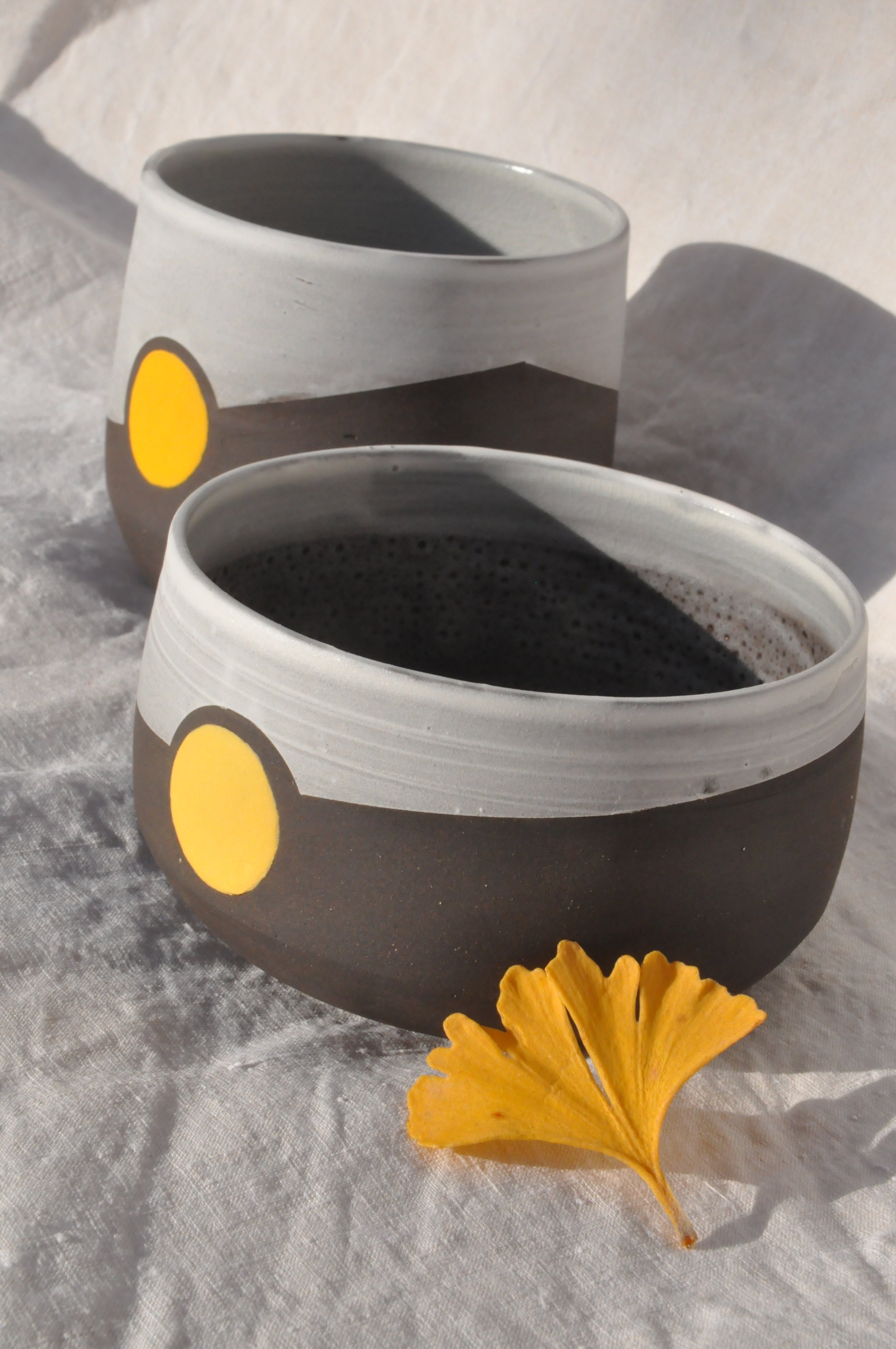Deux tasses à café en céramique, l'une remplie de café noir, posées sur une surface en tissu blanc. Une feuille d'érable jaune est placée devant la tasse en premier plan, avec une décoration de fleurs jaunes en dessous.