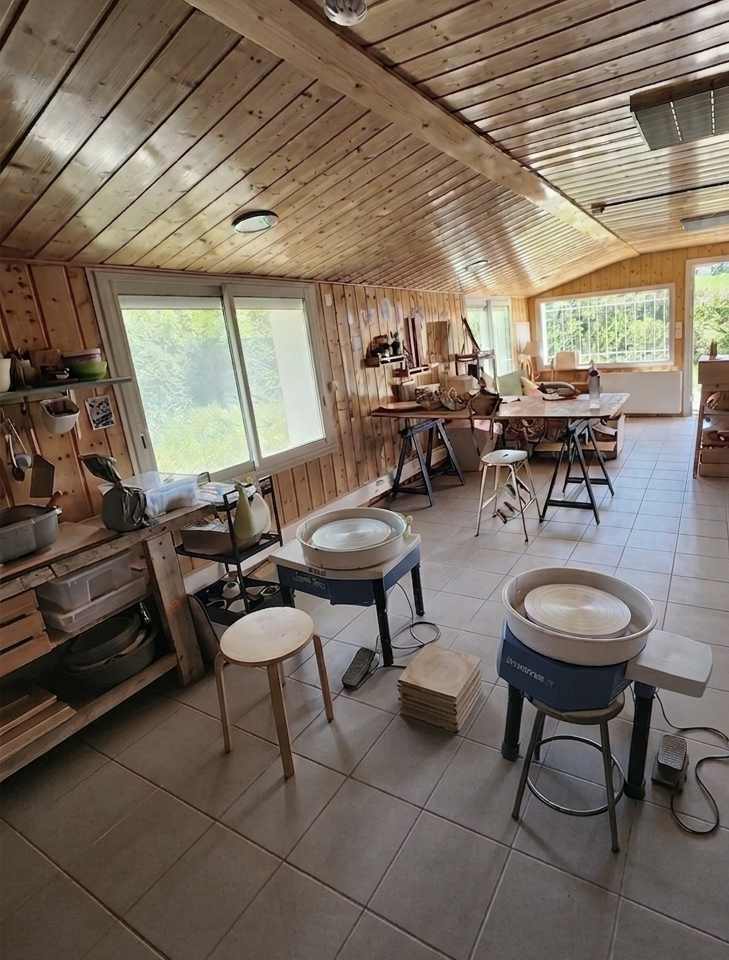 Une pièce en bois avec des fenêtres, un espace de travail pour la poterie avec des tours de poterie, des meubles en bois, et des matériaux de céramique.