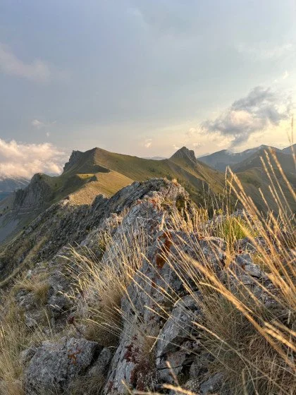 Paysage de montagne avec des collines verdoyantes, des rochers et de l'herbe hautes, sous un ciel partiellement nuageux au coucher du soleil.