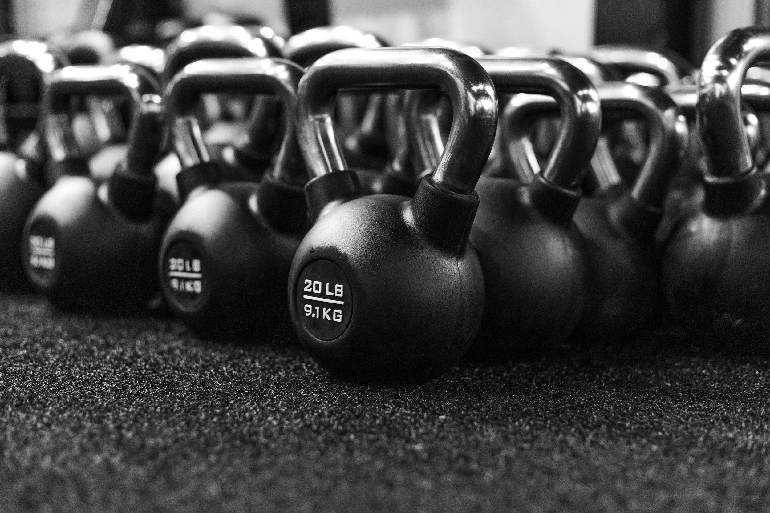 Black and white photograph of multiple kettlebells arranged in rows, labeled with weight in pounds and kilograms, on a textured surface.