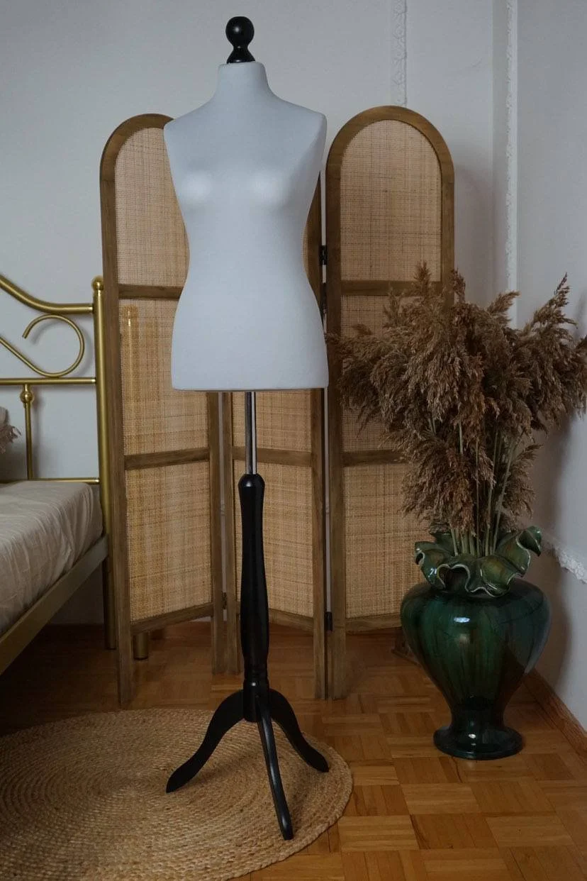 Eine Schneiderpuppe auf einem schwarzen Ständer, mit einer dekorativen Raumtrennwand, einer großen grünen Vase mit getrockneten Pflanzen und einem Holzfussboden.