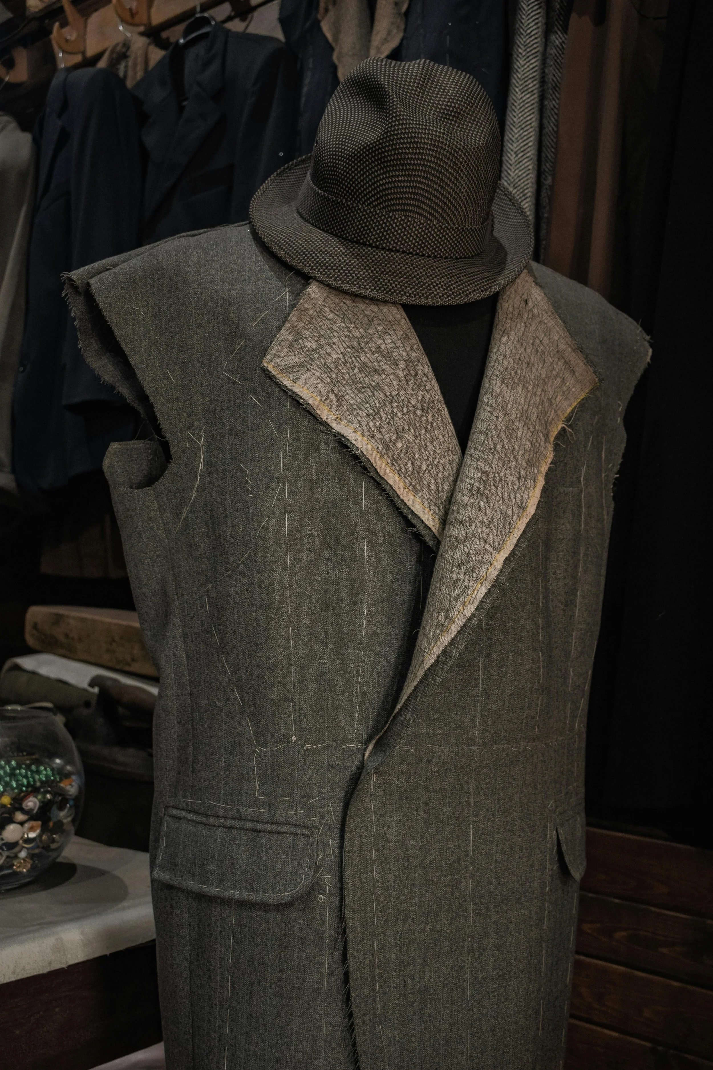 Mannequin mit einem grauen Wollanzug und einem schwarzen Hut, im Hintergrund sind verschiedene Jacken und Mäntel aufgehangen.