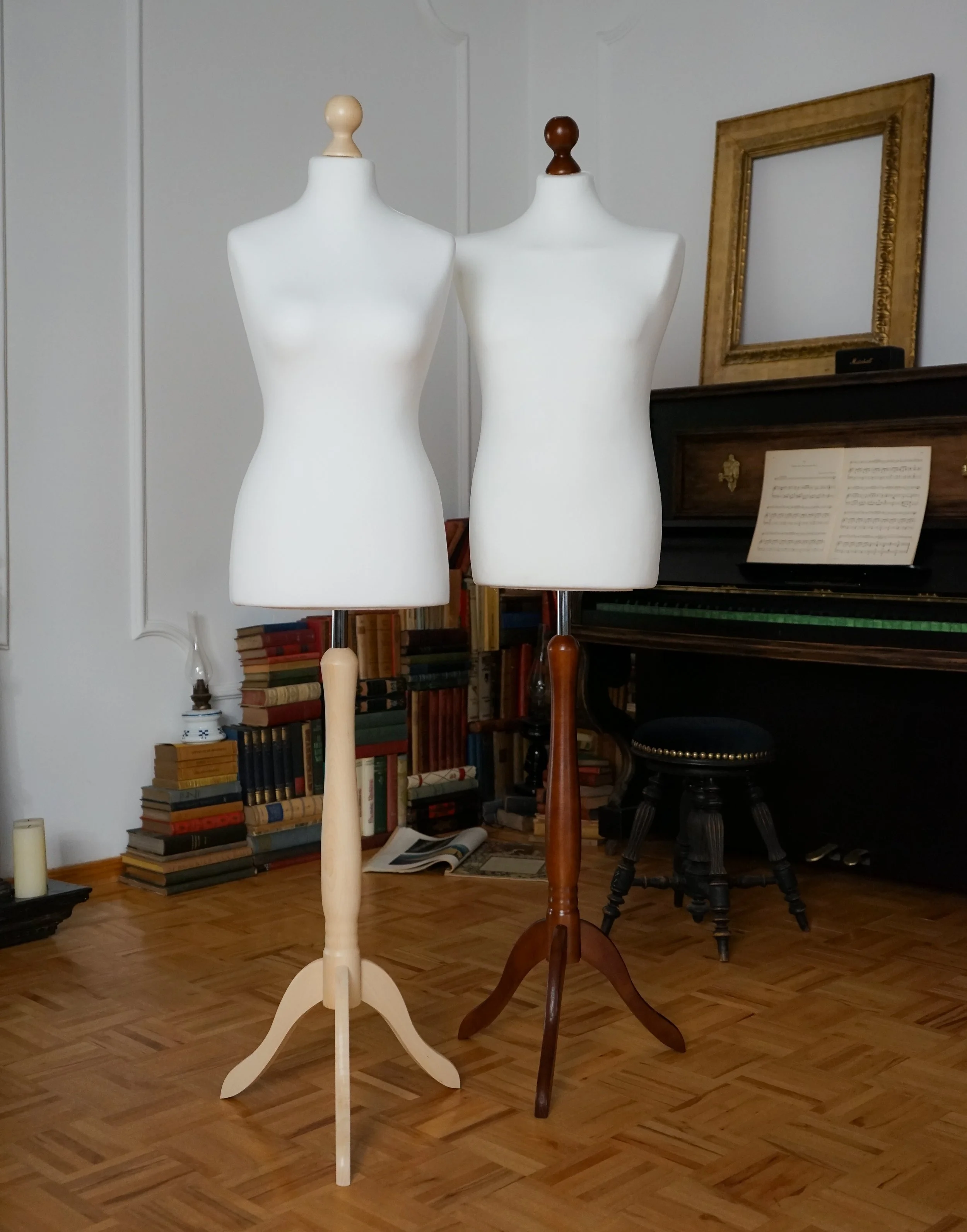 Mannequins de couture en bois, blancs, placés dans une pièce avec une bibliothèque, un piano avec une partition de musique et une chaise à pied en bois.