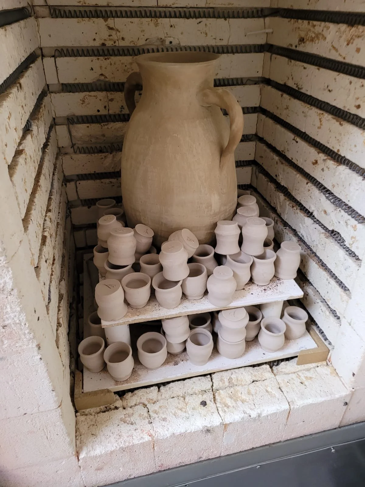 Onbewerkte keramische pot en kleine keramieke potten op een houten plank in een bakkerij of pottenbakkerij, met bakstenen muur en metalen draden.