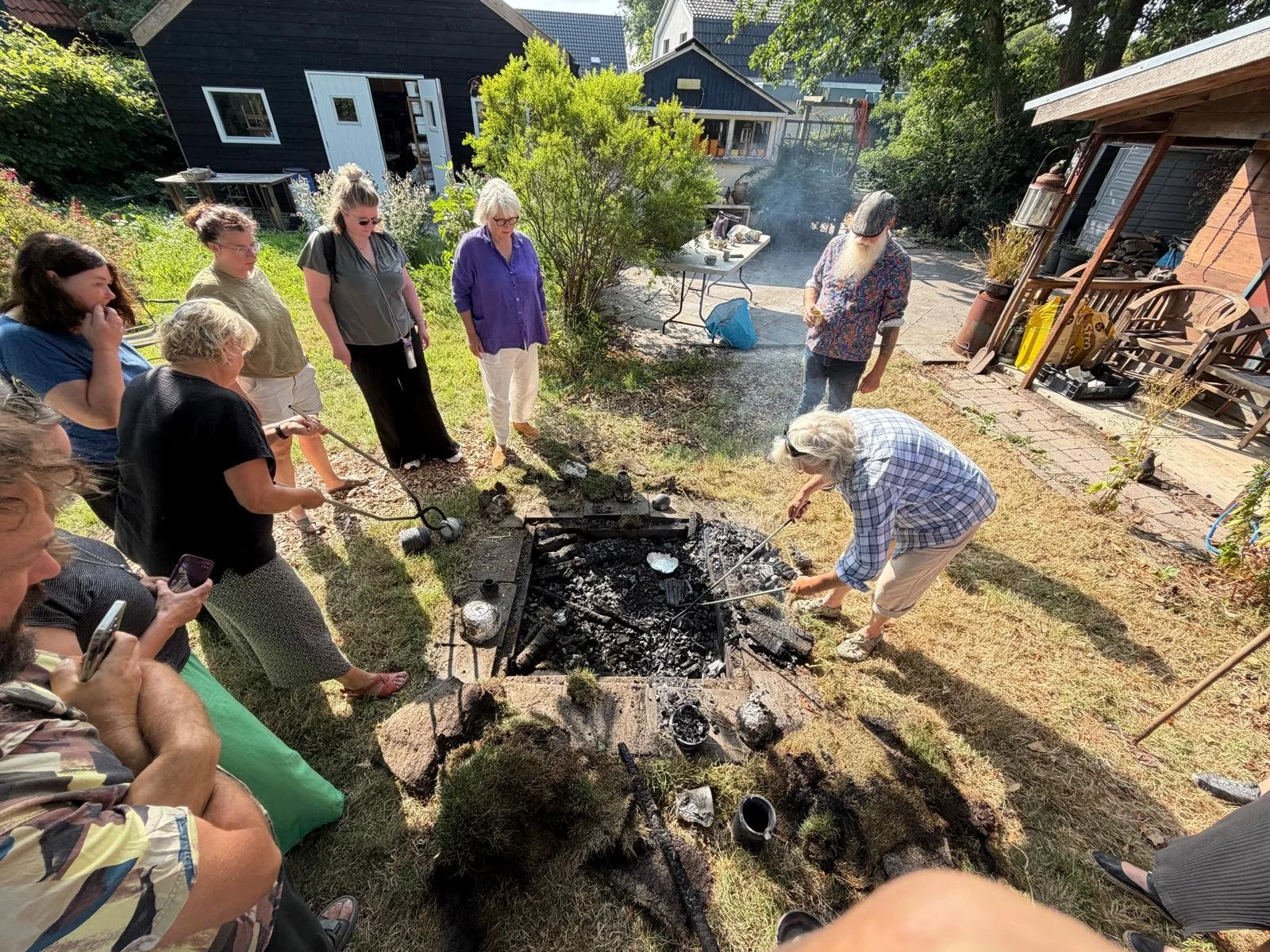 Mensen verzamelen zich rondom een tuinplaats met een kampvuur, waar twee mensen hout bijstoken. Sommigen maken foto's of kijken toe, in een zonnige tuin met bomen en schuren op de achtergrond.