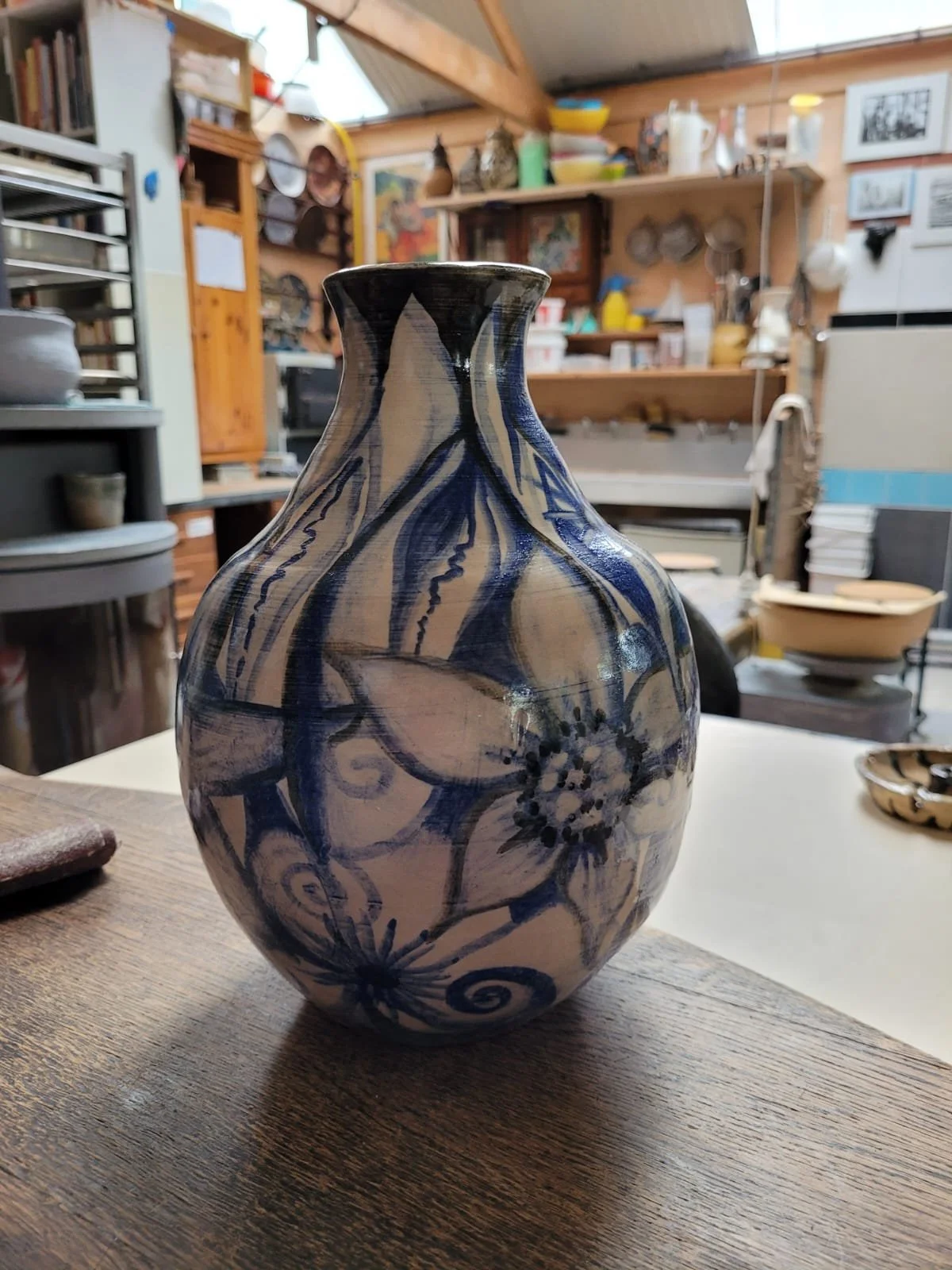 Een keramische vaas met blauwe bloem- en krabbelpatronen op een houten tafel in een atelier.
