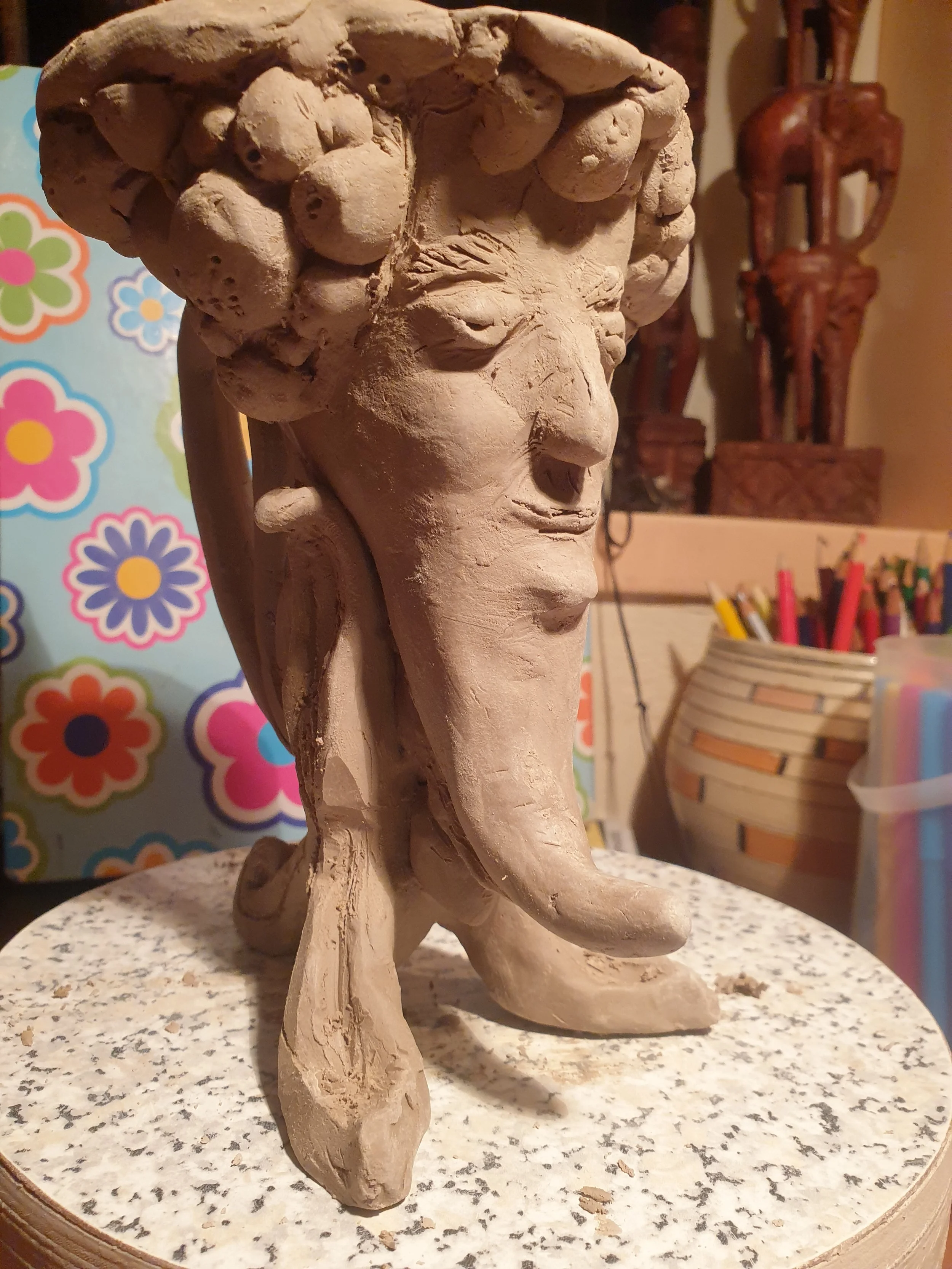 Een onafgewerkte beeldhouwwerk van een menselijk gezicht met complexe haarstructuur, gemaakt van klei, geplaatst op een ronde tafel in een atelier.