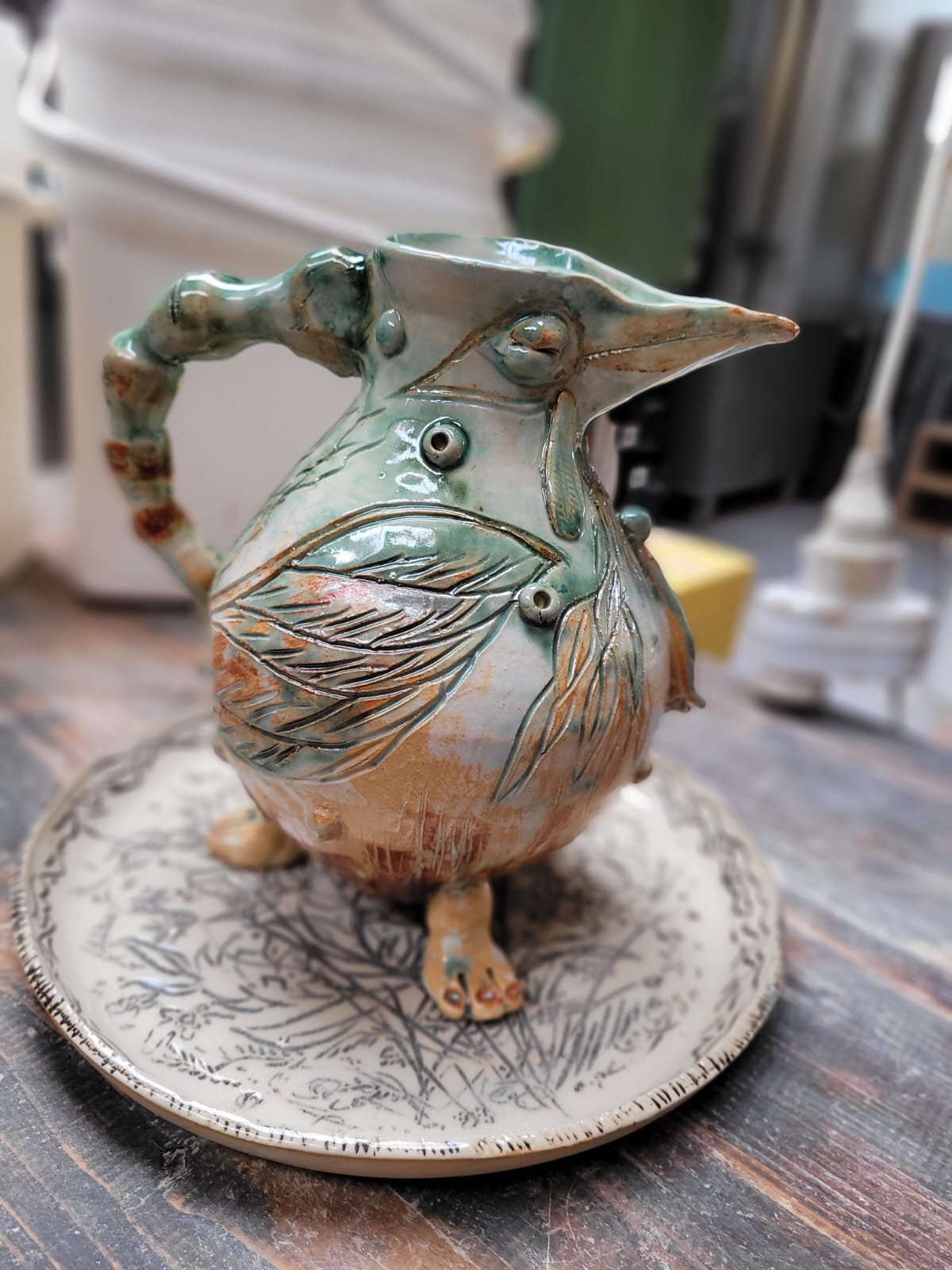 Een keramieken vaas in de vorm van een vogel met details van een blad op de lichaam en een gebogen snavel. De vaas staat op een ronde schaal met een bladpatroon.