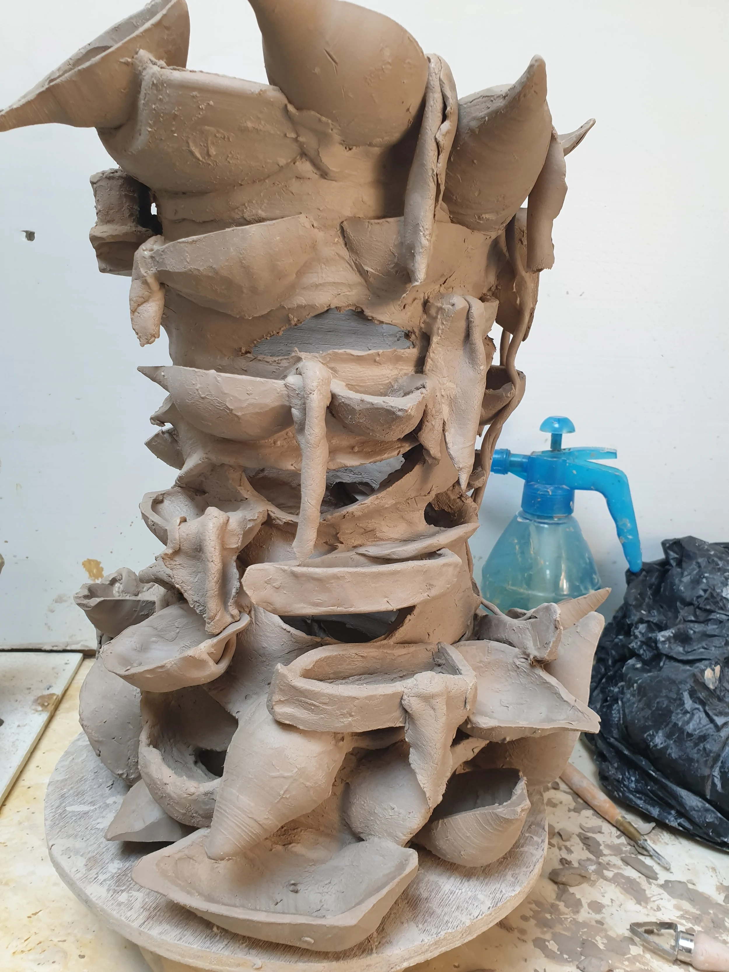Onbewerkt keramisch beeldhouwwerk met meerdere natuurlijke vormen en uitsparingen, op een draaischijf, omgeven door gereedschap en een blauw sproeiflesje.