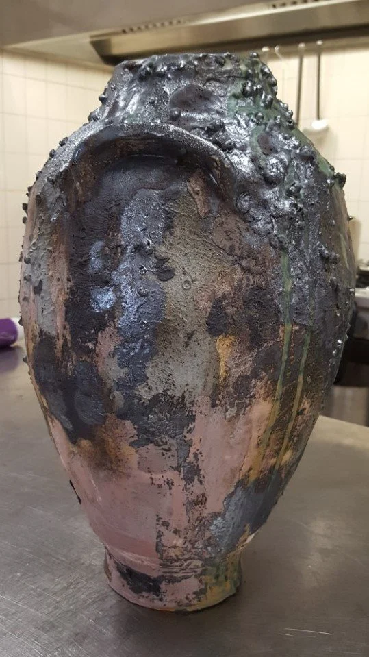 Een vaas met een onregelmatige, ruwe en gekleurde oppervlakte, mogelijk keramiek of gestookte kunst. Het ziet eruit als een kunstwerk of decoratief voorwerp.