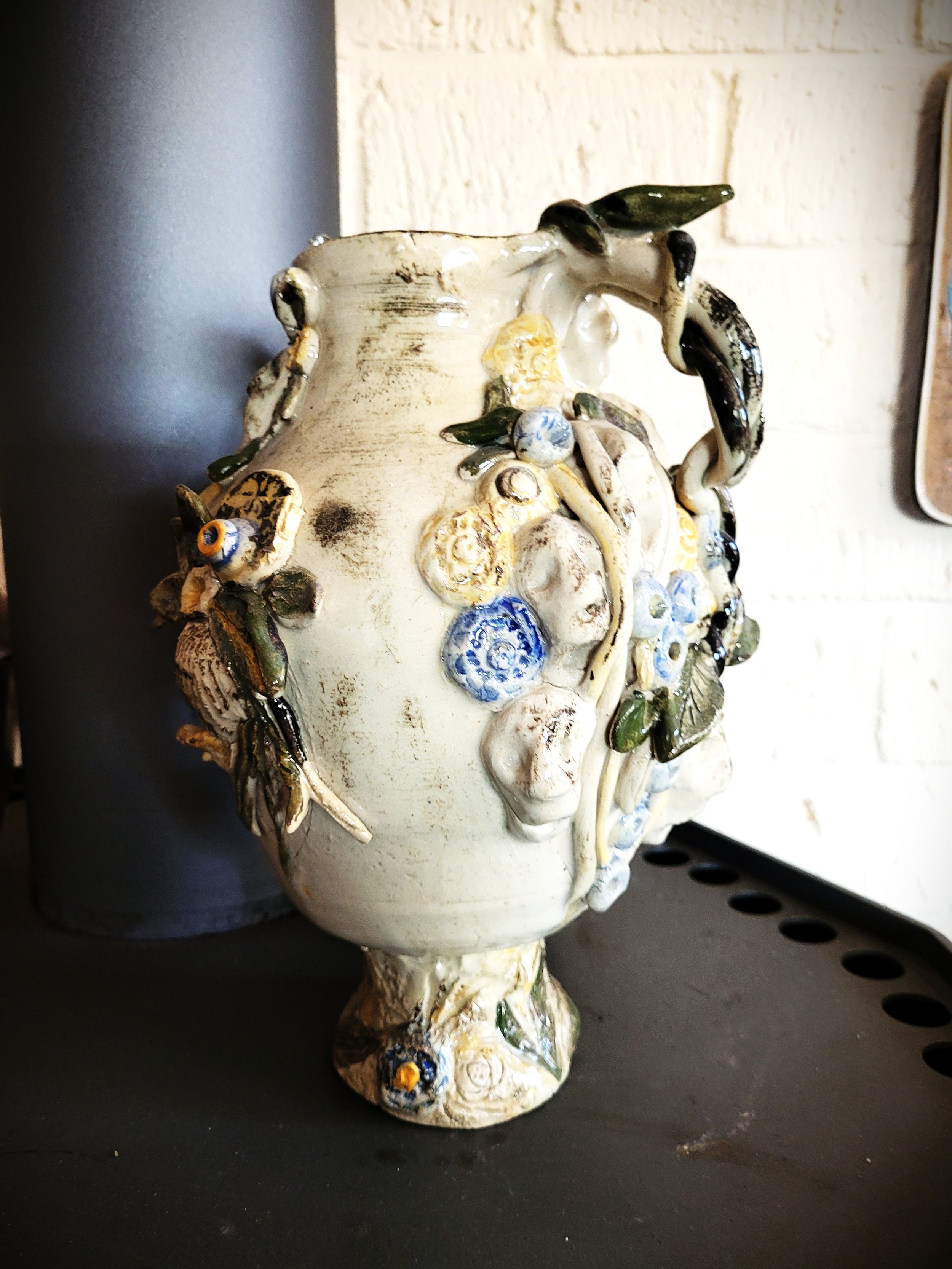 Een sierpot met een gedetailleerd reliëf van bloemen en bladeren, vervaardigd uit keramiek of porselein, geplaatst op een zwarte ondergrond en tegen een witte bakstenen muur.