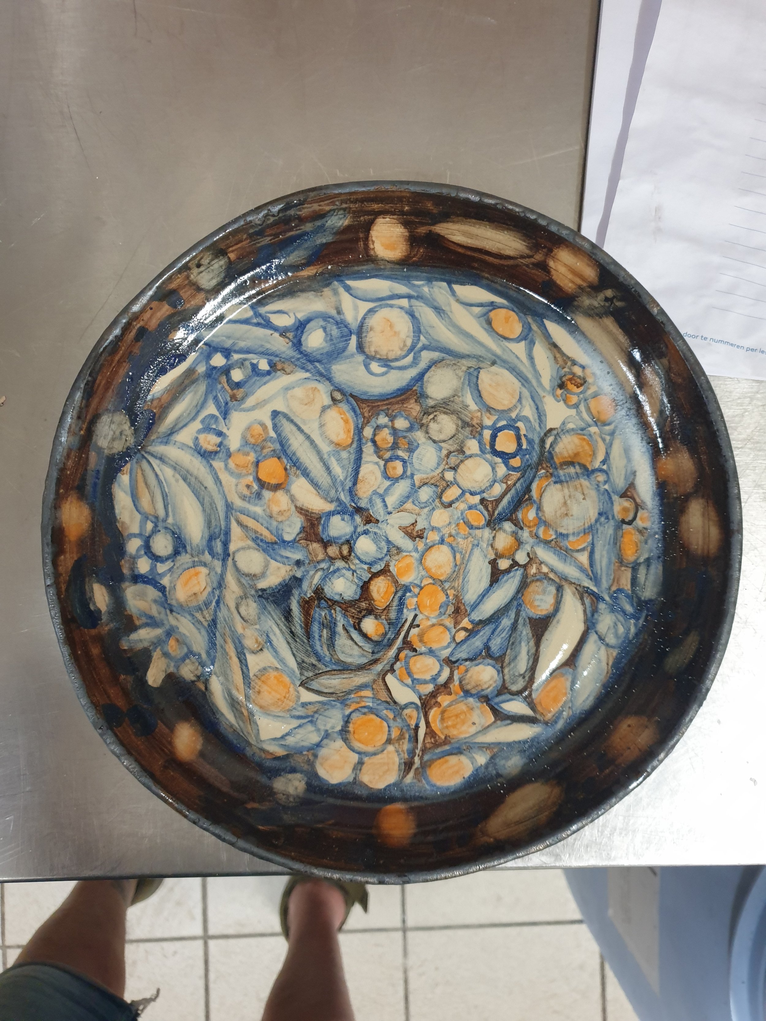 Een keramische schaal met een blauw en oranje decoratief patroon, geplaatst op een metalen tafel, met de voeten van een persoon eronder zichtbaar.