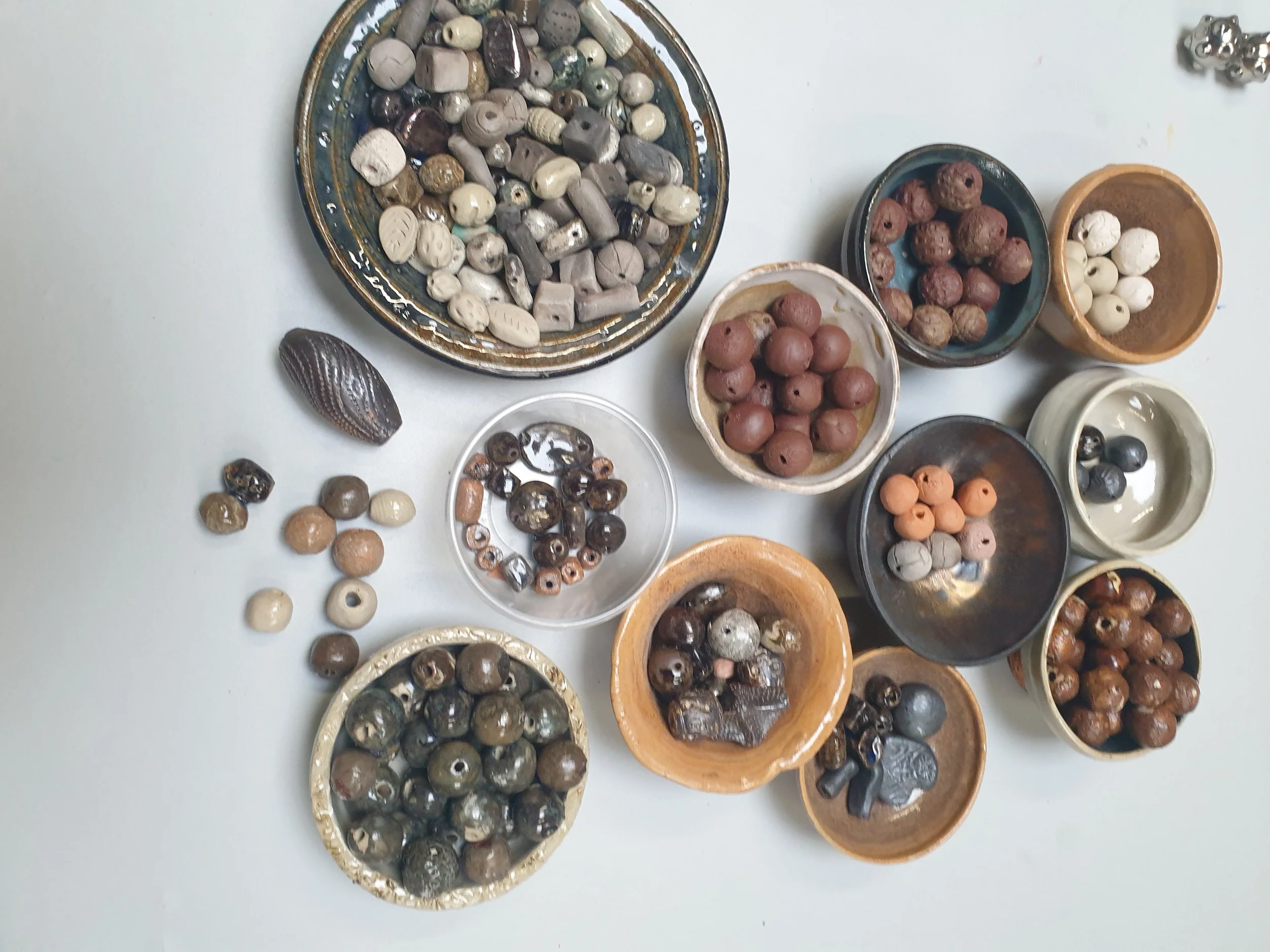 Verschillende soorten kralen en zaden in kleine potjes en op een bord, variërend in kleur en vorm.