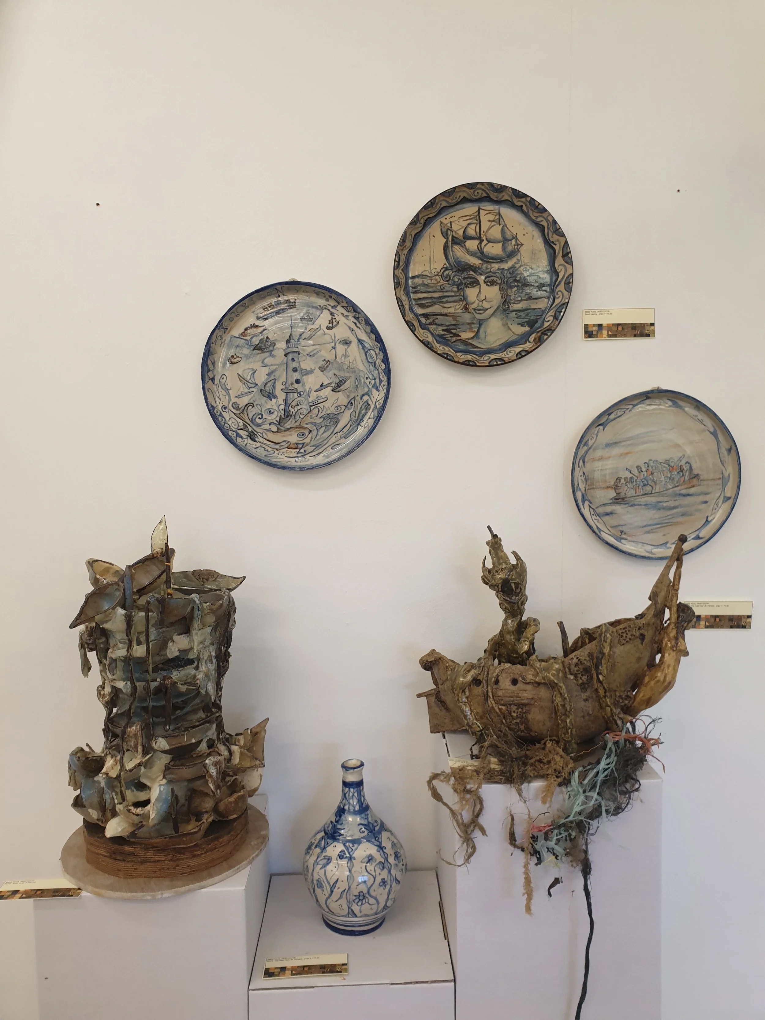 Twee blauwe en witte gerijpte aardewerkschalen met schelpmotieven, drie sculpturale kunstwerken van hout en natuurlijke materialen, en twee glazen vazen met blauw en wit decor, allemaal tentoongesteld in een museum of galerij.