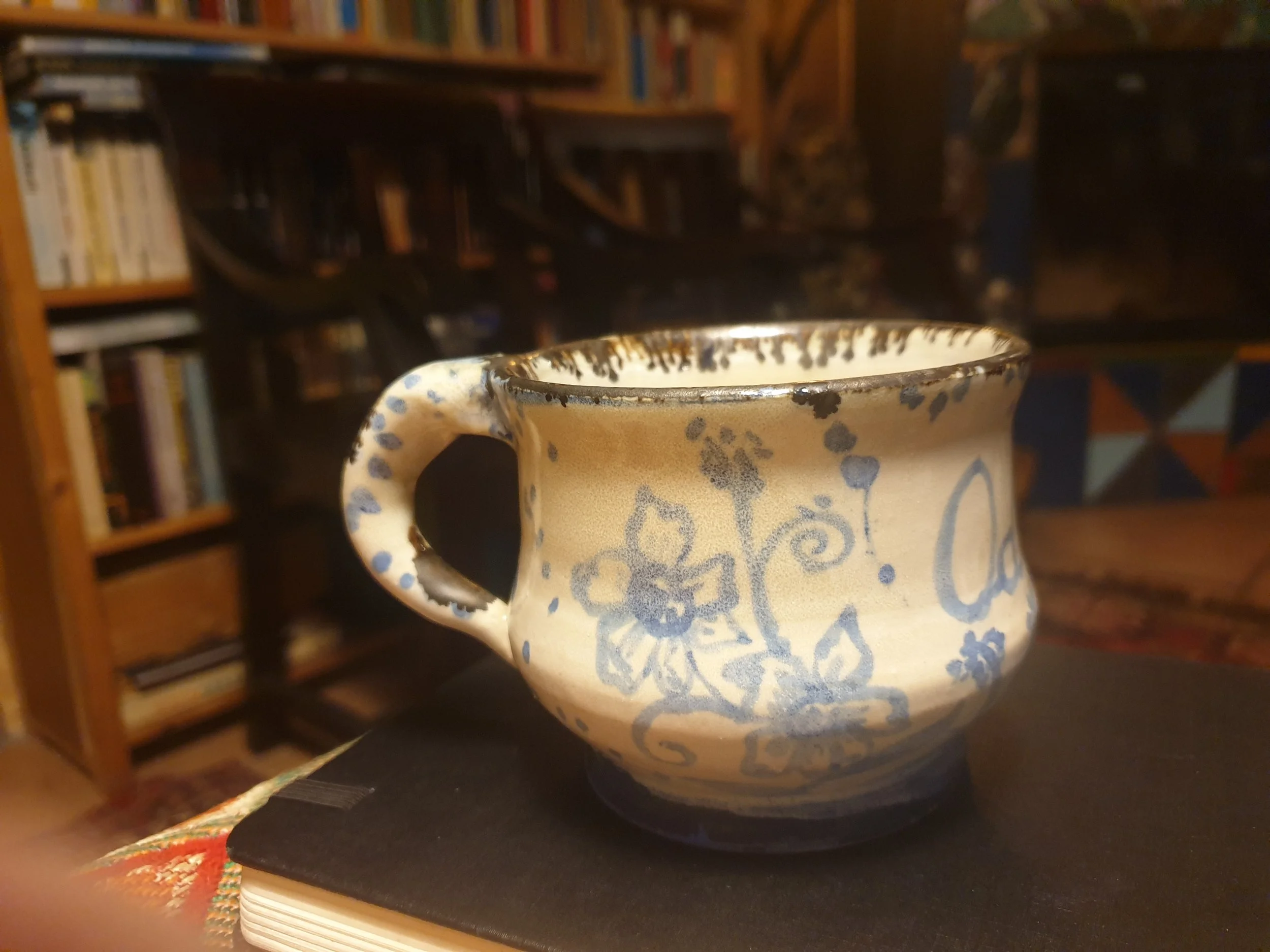 Een keramieke mok met blauwe patronen vooraan op een boekenplank, op de achtergrond boeken.