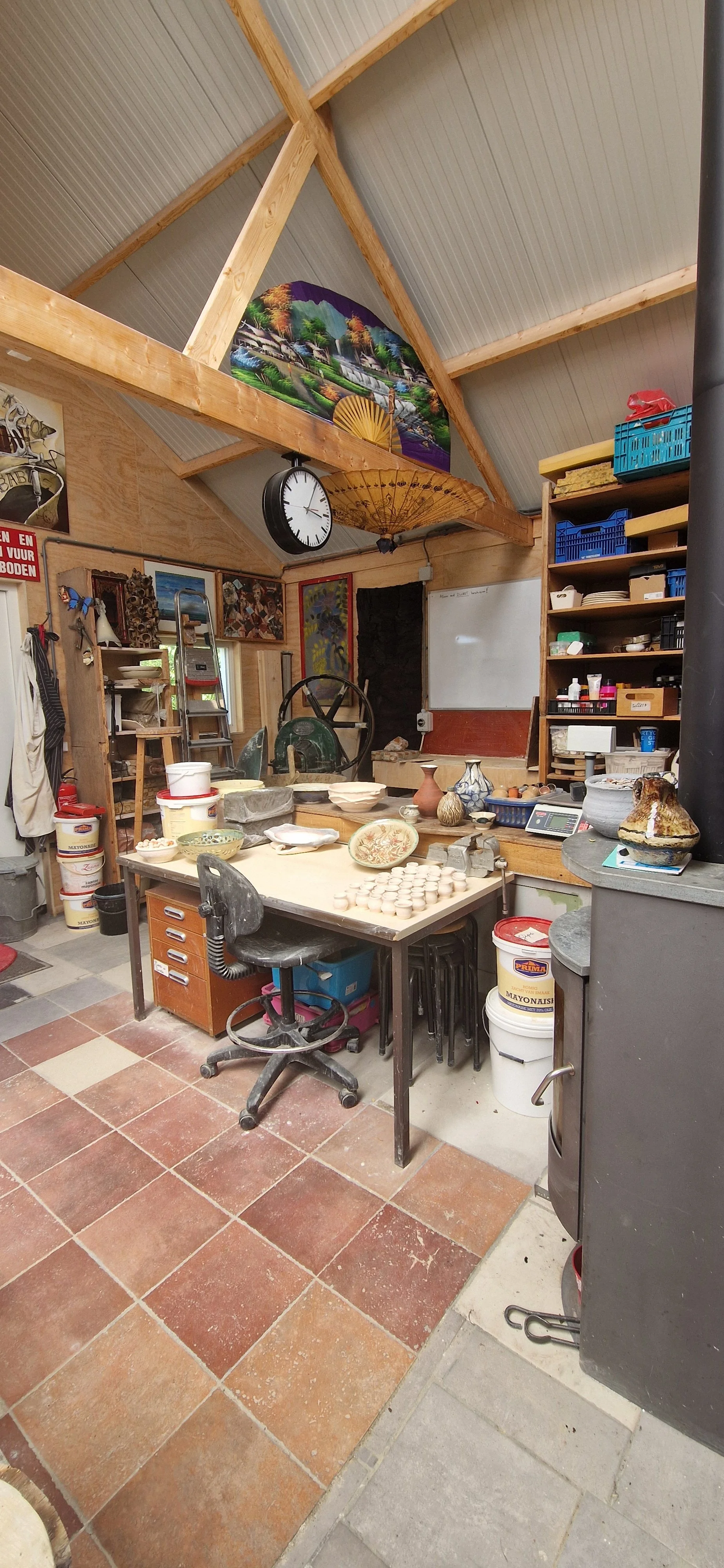 Een werkplaats met een tafel vol keramische werkstukken en potten, omringd door gereedschap, opslagrekken en decoratieve kunstwerken.