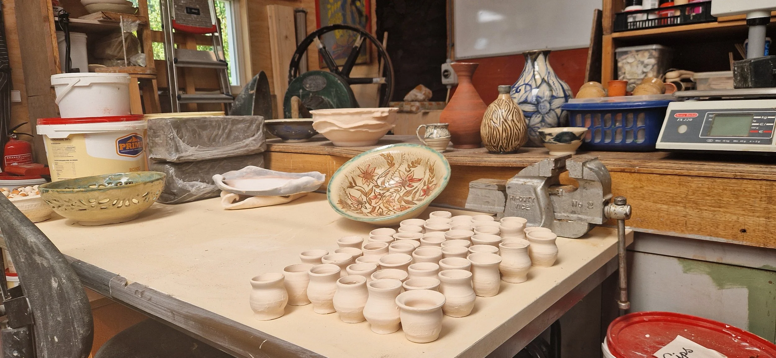 Tafel met verschillende keramieke potten, kommen, en vazen in een pottenbakkerij, met gereedschap en apparatuur op de werkplek.