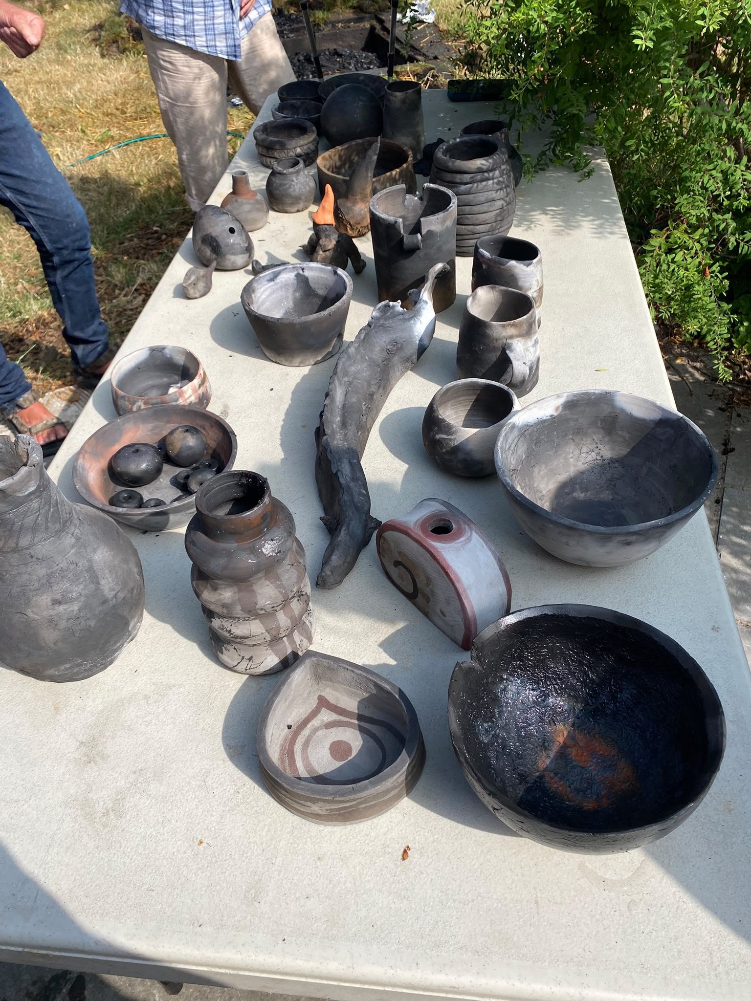 Een buitentafel met diverse keramieken vazen en potten, waarschijnlijk handgemaakt, in verschillende vormen en kleuren, met mensen op de achtergrond.