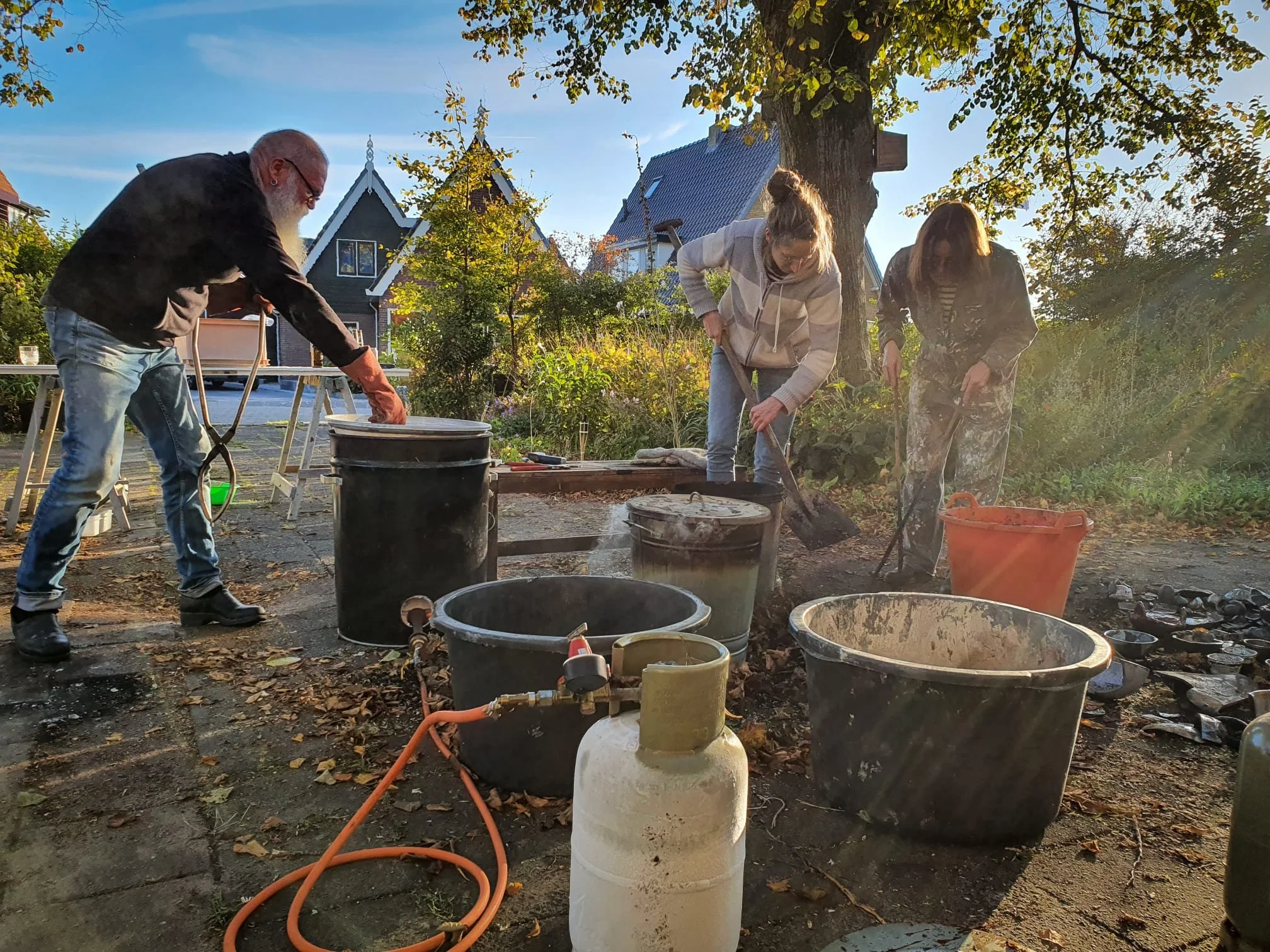 Drie mensen werken samen in een tuin, bezig met het plantwerk. Ze staan naast een grote boom, omringd door verschillende containers en tuinmaterialen, terwijl de zon ondergaat.