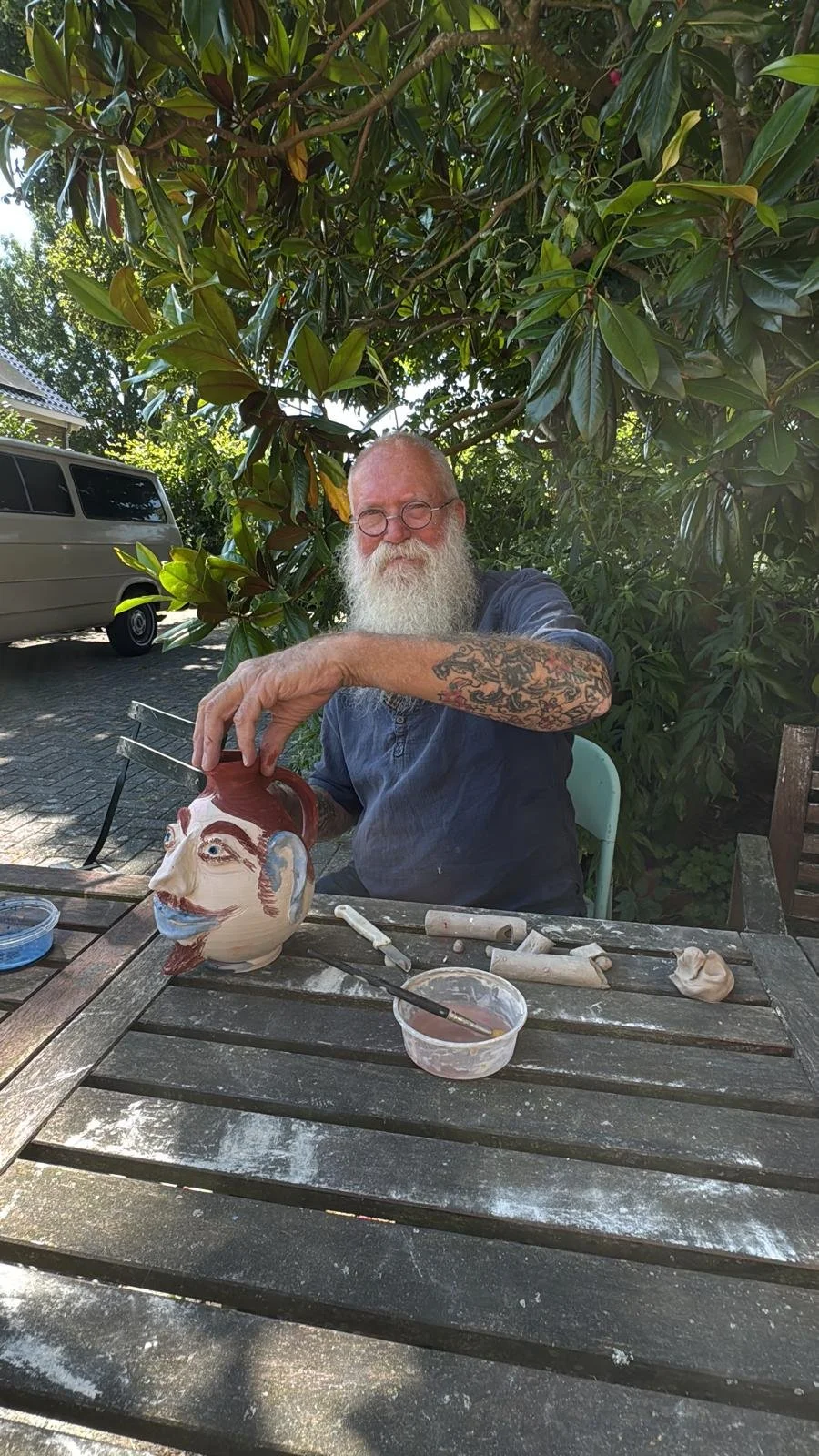 Kunstenaar Baba werkt aan een grote keramische vaas met een gezicht op een houten tafel buiten, onder een boom.