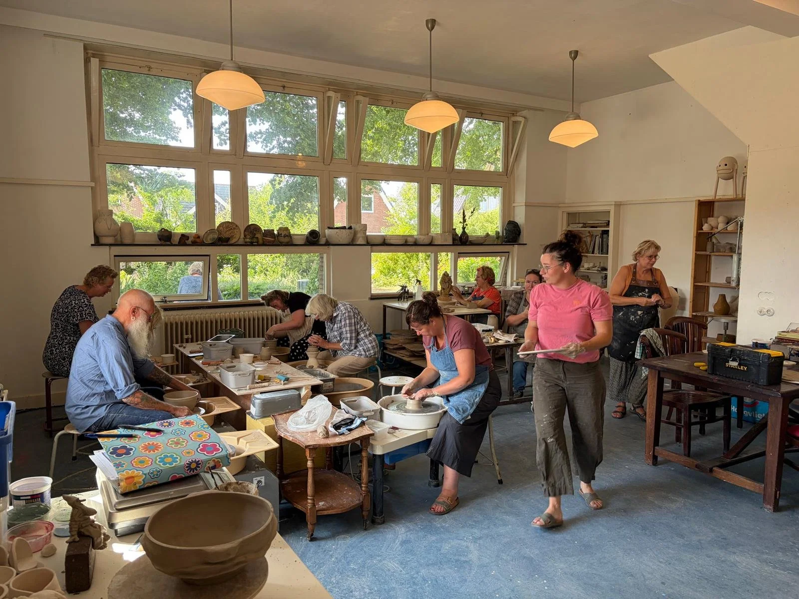Mensen werken aan verschillende keramiekprojecten in een licht en ruim atelier met grote ramen en groene bomen buiten.