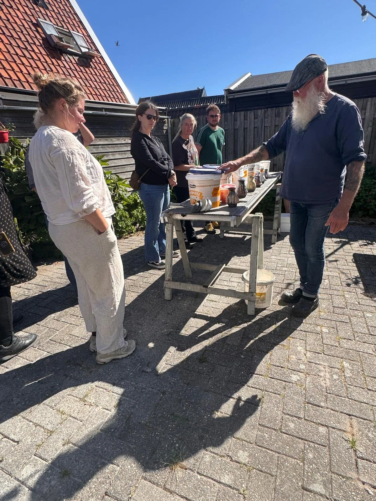 Een groep mensen staat buiten op een zonnige dag rond een tafel met potjes en bekers, terwijl een man met een grijze baard en pet iets uitlegt.