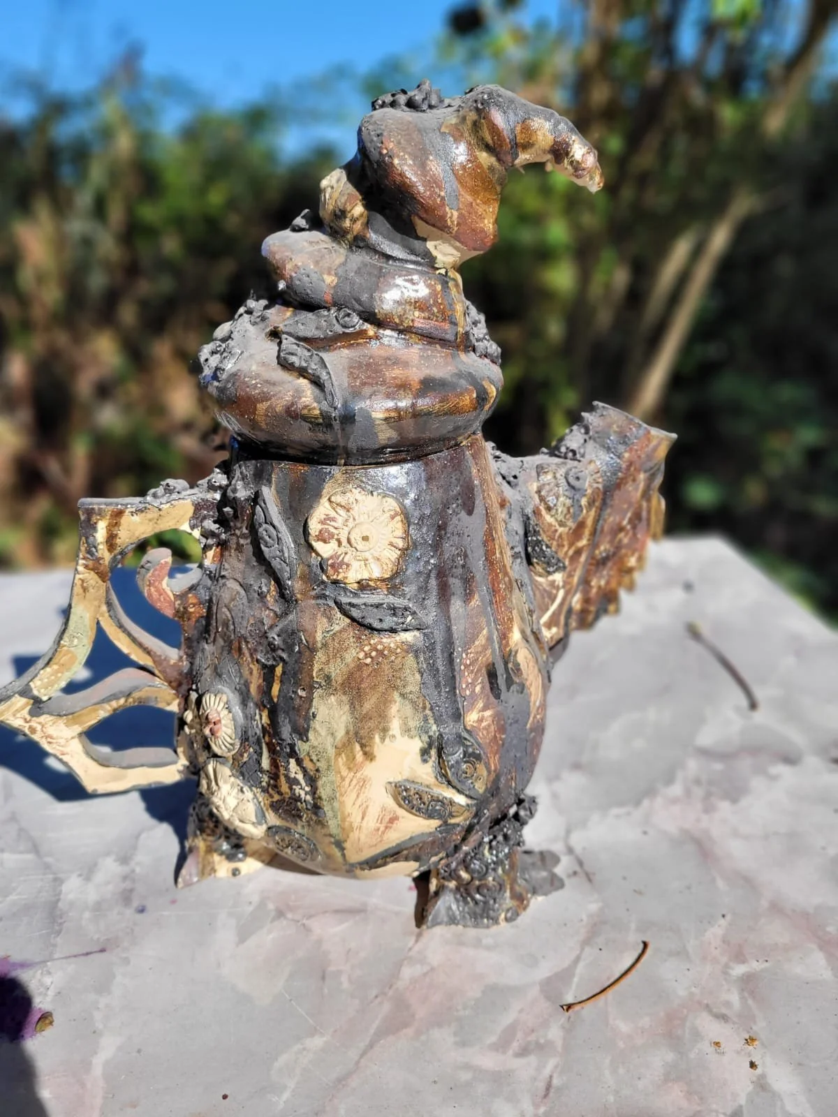 Antieke metalen theepot met gedetailleerde decoraties en reliëfs, op een houten tafel buiten met een groene boom en blauwe lucht op de achtergrond.