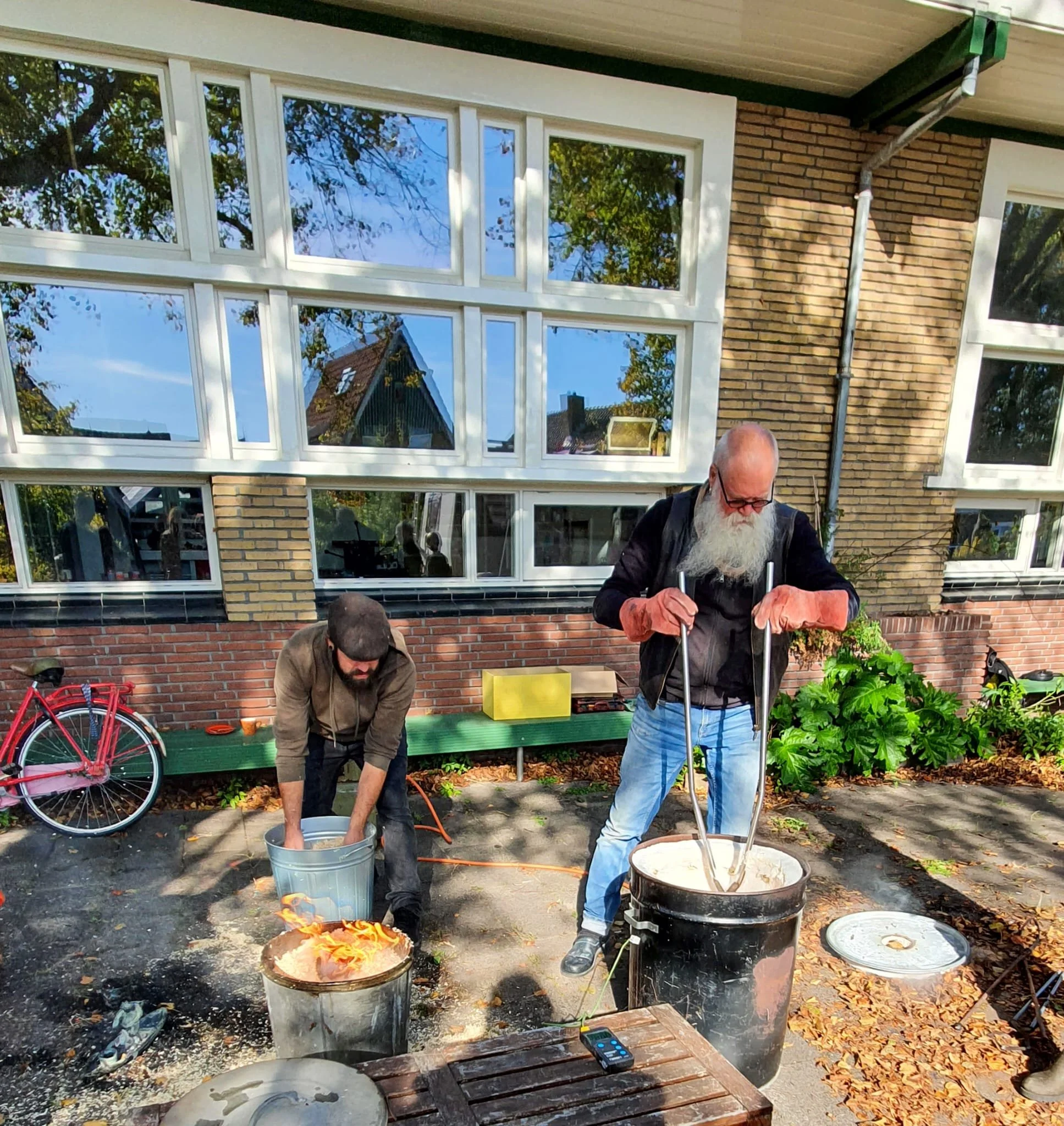 Twee mannen in een tuin, bezig met een brandende container en smeltende kaarsen, met een huis met grote ramen en een tuin vol planten op de achtergrond.