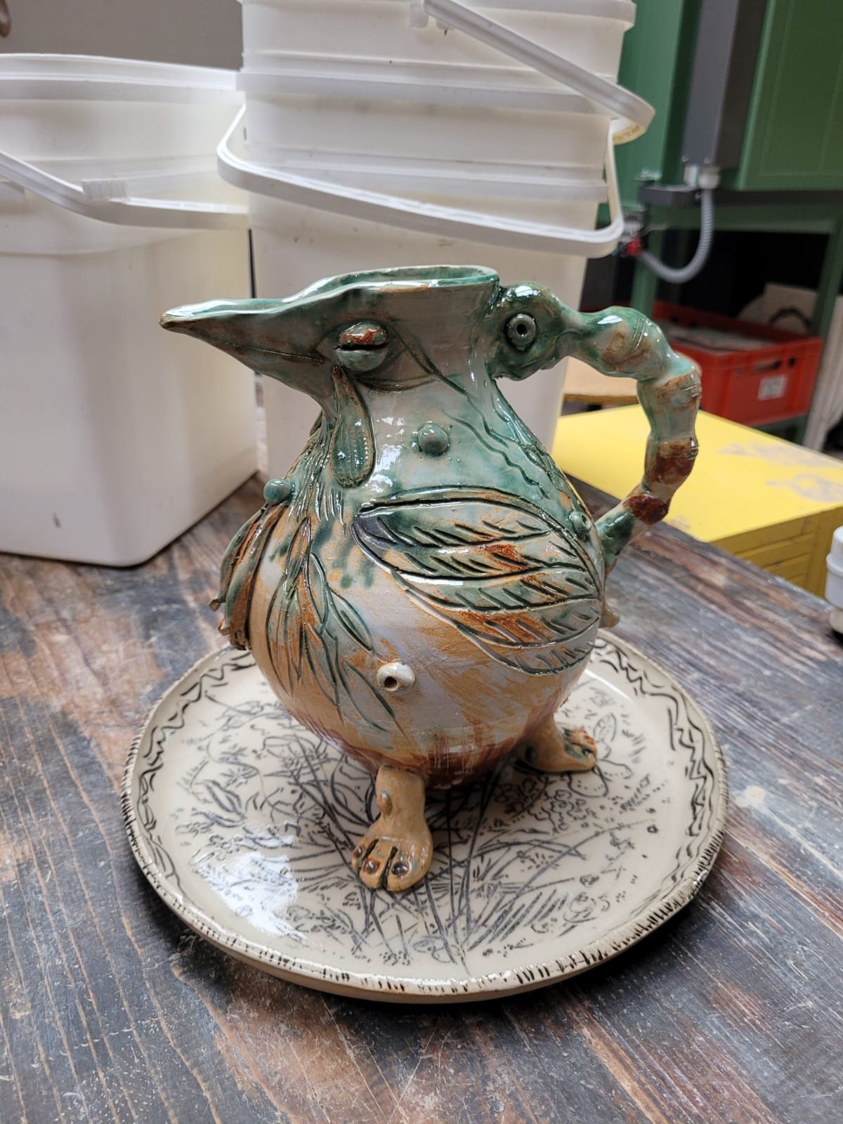 Een keramieken thee- of waterkruik in de vorm van een dier met een grote buik, een lange snuit, en een handvat, versierd met bladeren en natuurlijke kleuren, geplaatst op een ronde dienblad op een houten tafel.
