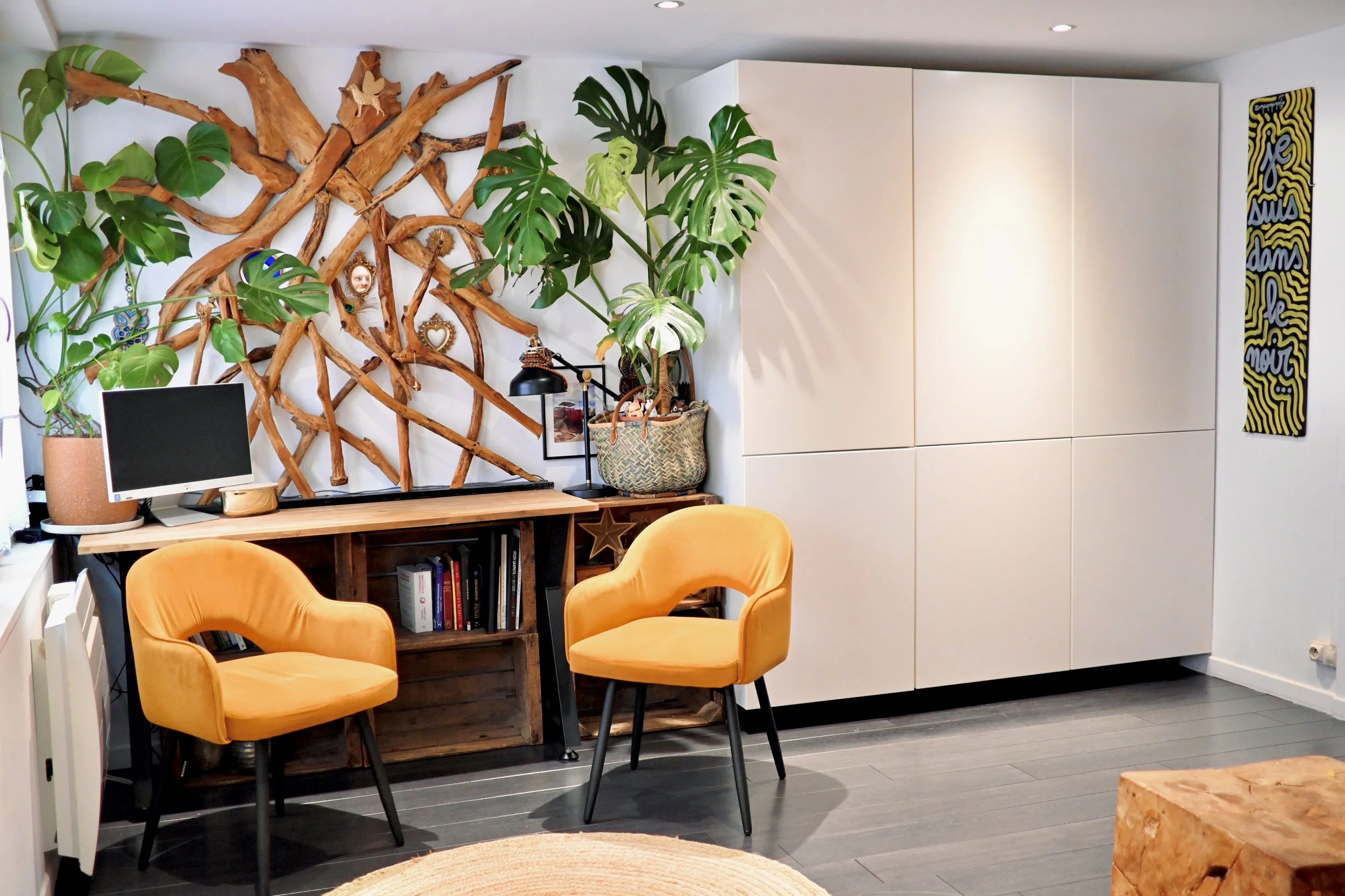 Un intérieur moderne avec des fauteuils orange, un bureau en bois, des plantes vertes, une armoire blanche, un tapis, et une décoration murale colorée.