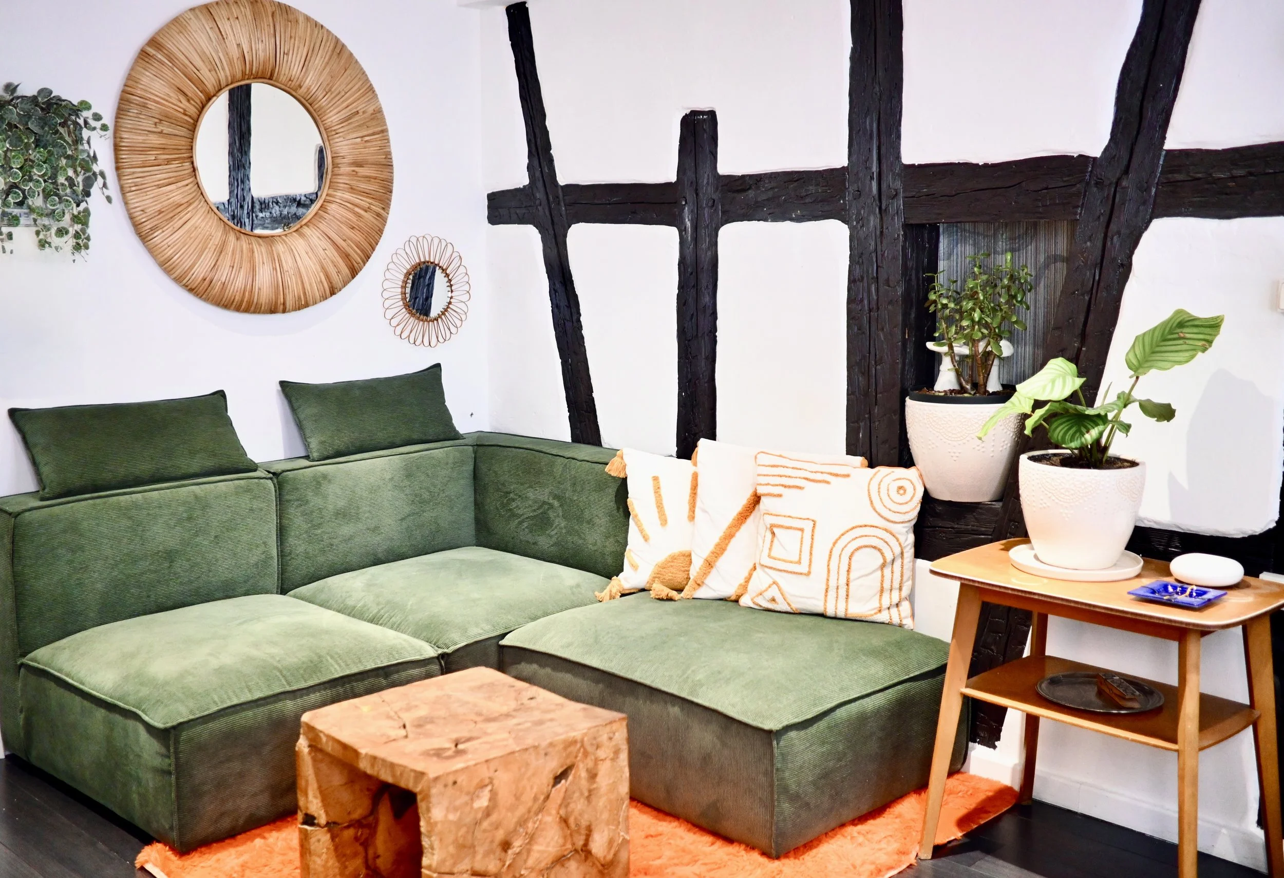 Un coin salon avec un canapé vert, plusieurs coussins, une table en bois et des plantes dans des pots blancs. Sur le mur, un miroir rond en rotin, un petit miroir décoratif et des poutres en bois noir.