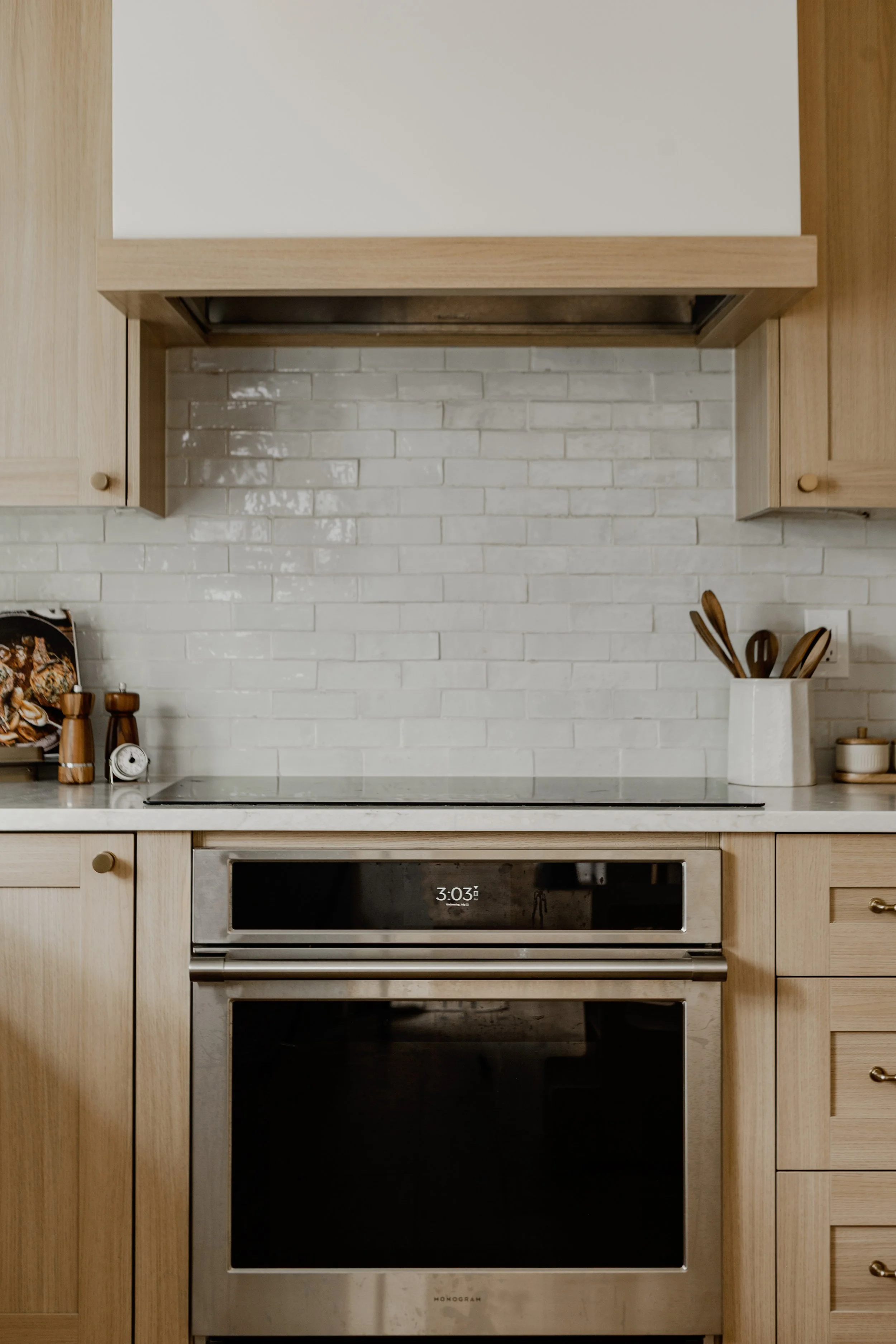 A kitchen with wooden cabinets, a white brick backsplash, and a stainless steel oven with a digital clock showing 3:03.