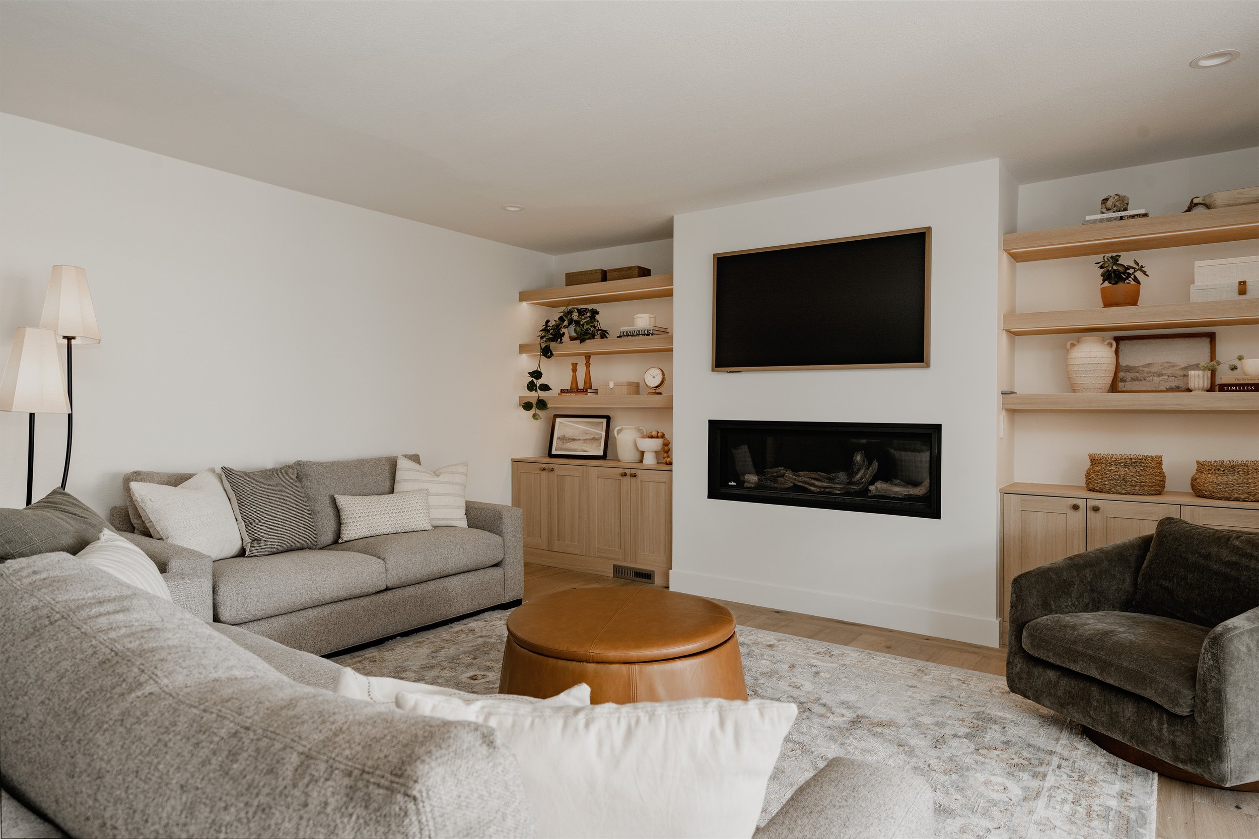 Living room with gray sofa, black armchair, wooden coffee table, beige and white pillows, white walls, wooden shelves with plants and books, wall-mounted flat-screen TV above a fireplace, and a floor lamp.