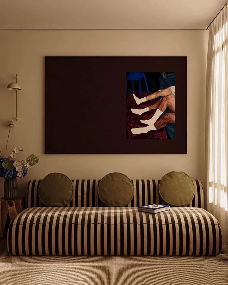 Living room with a striped sofa, three green pillows, a book on the sofa, a wall hanging with abstract artwork, a side table with flowers, and curtains letting in daylight.