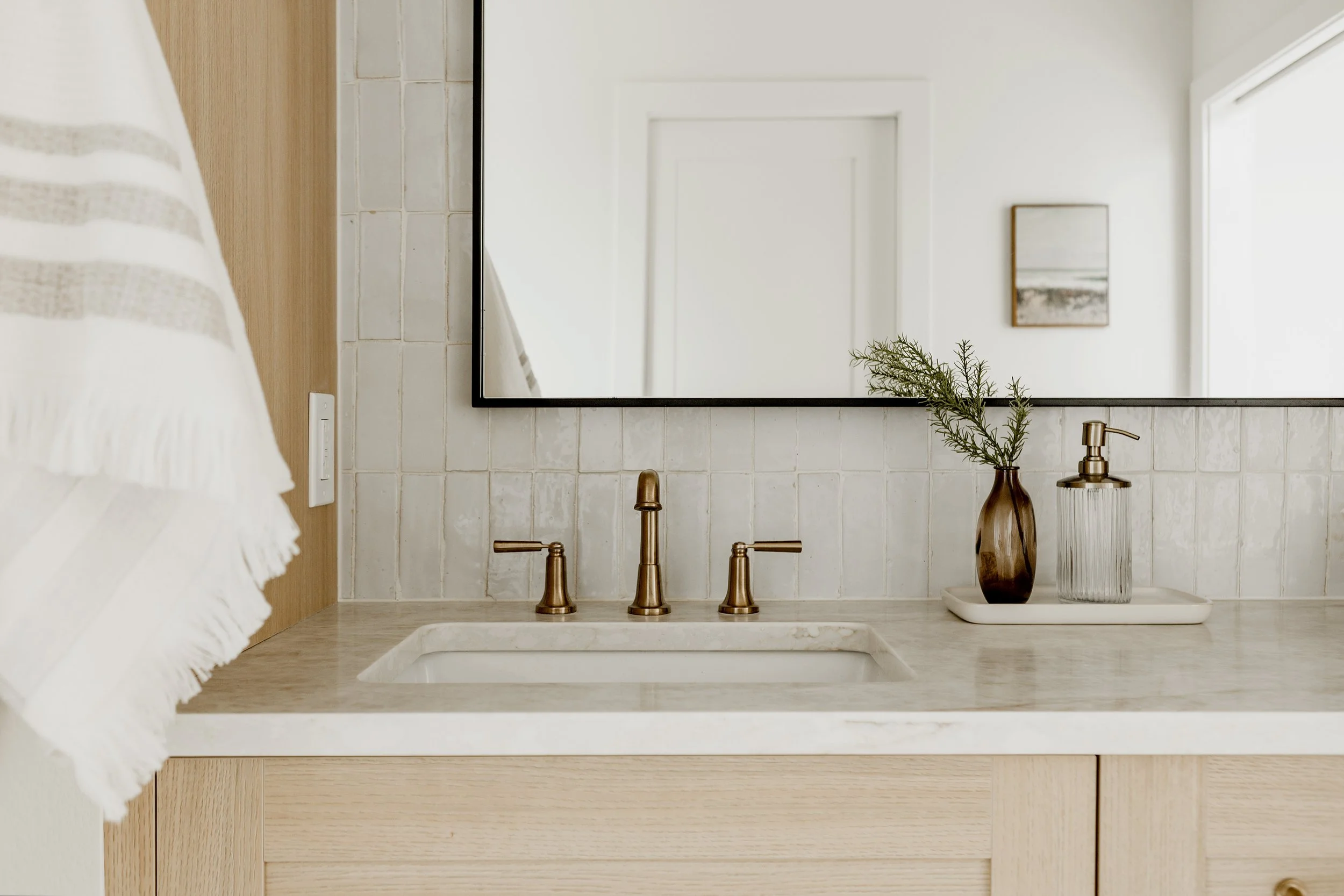 Bathroom sink with a gold faucet, a mirror, a framed artwork, and decorative vases with greenery.