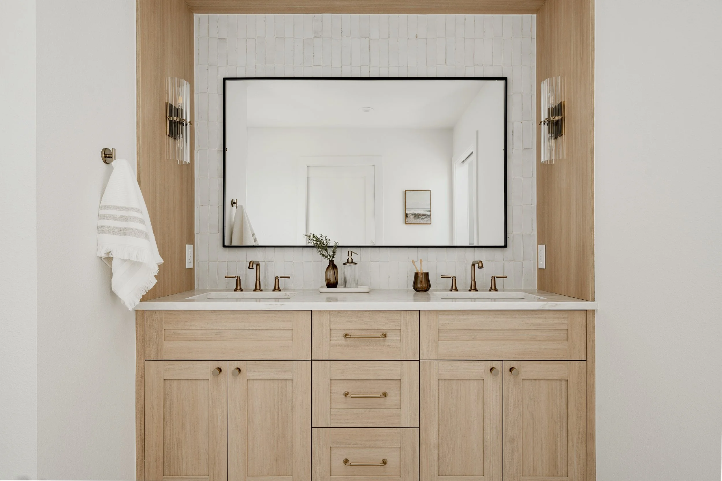 Bathroom vanity with two sinks, light wood cabinetry, large rectangular mirror, and wall-mounted light fixtures.