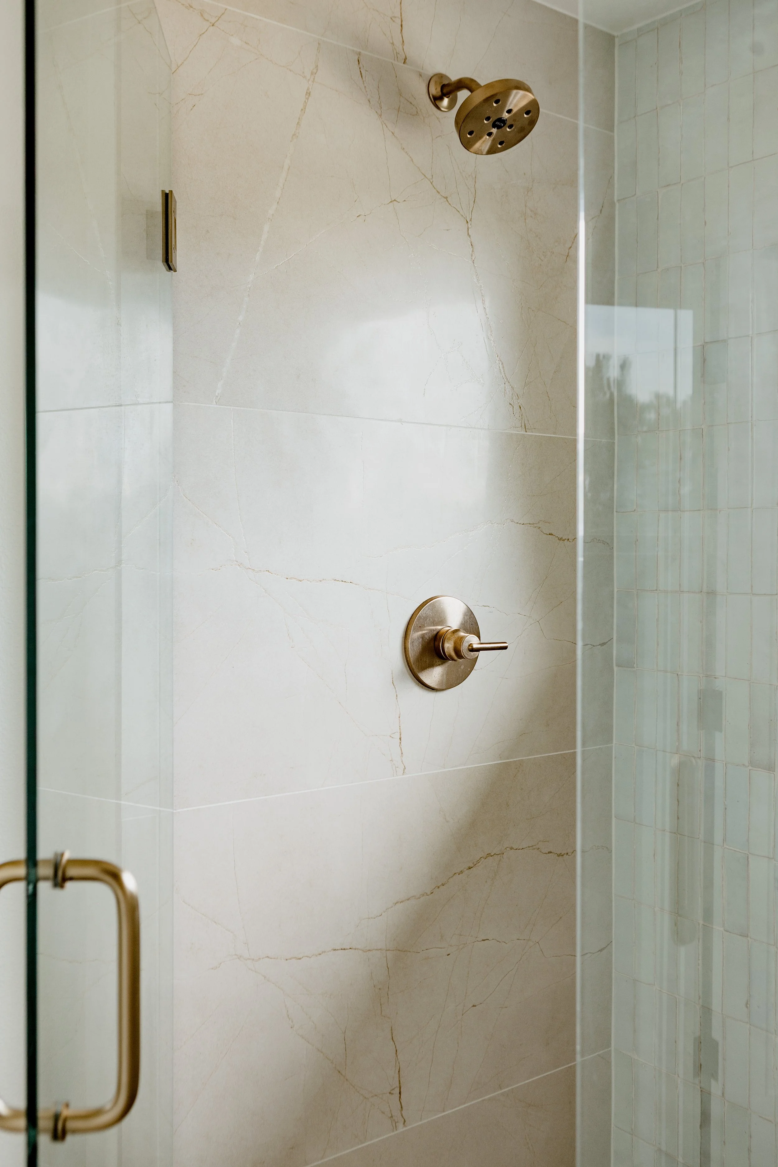 Shower with beige marble walls, a gold showerhead, a gold handle, and a glass door with a gold handle.