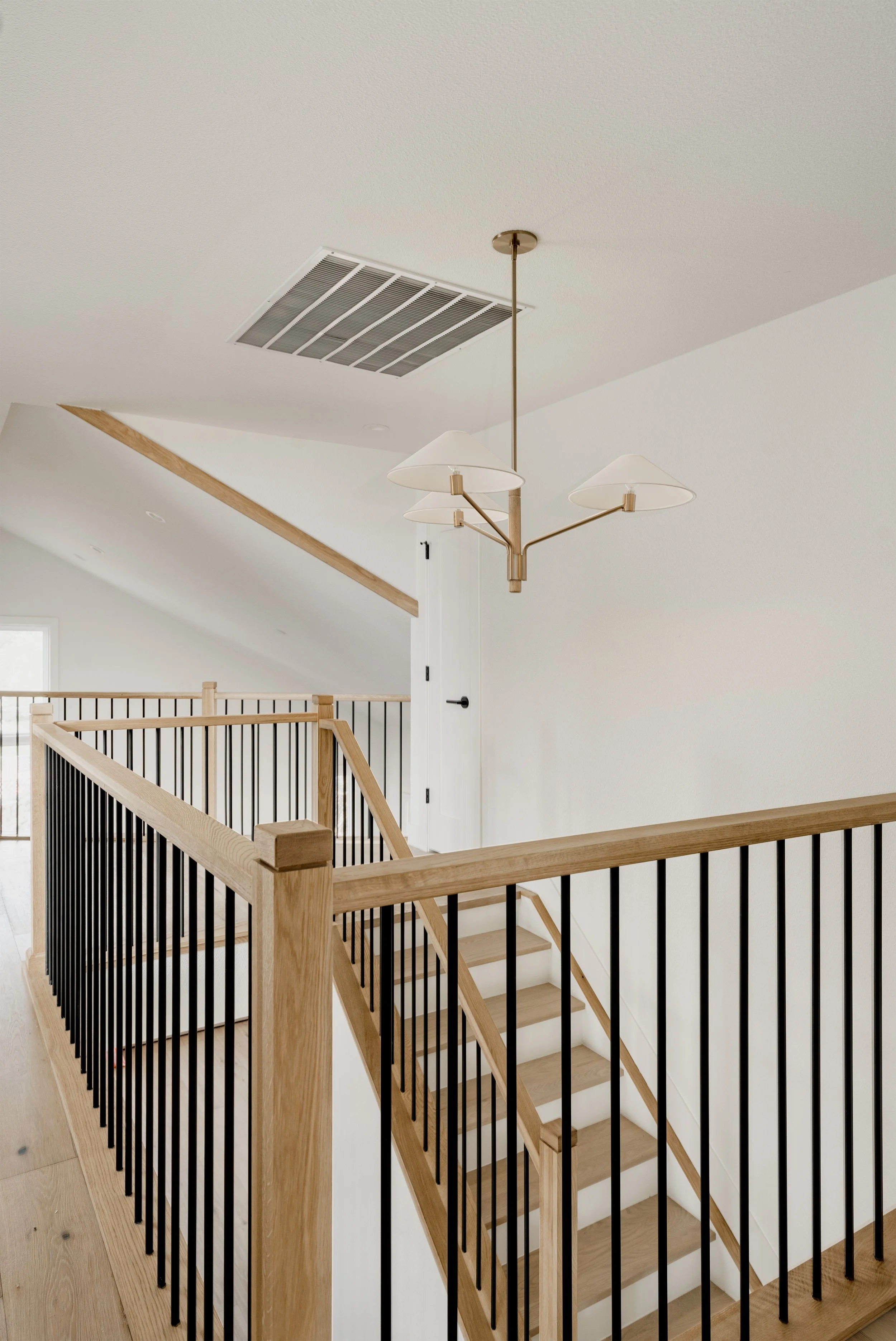 Interior view of a staircase with wooden handrail and black vertical balusters, white walls, and a modern chandelier with three white lampshades hanging from the ceiling.