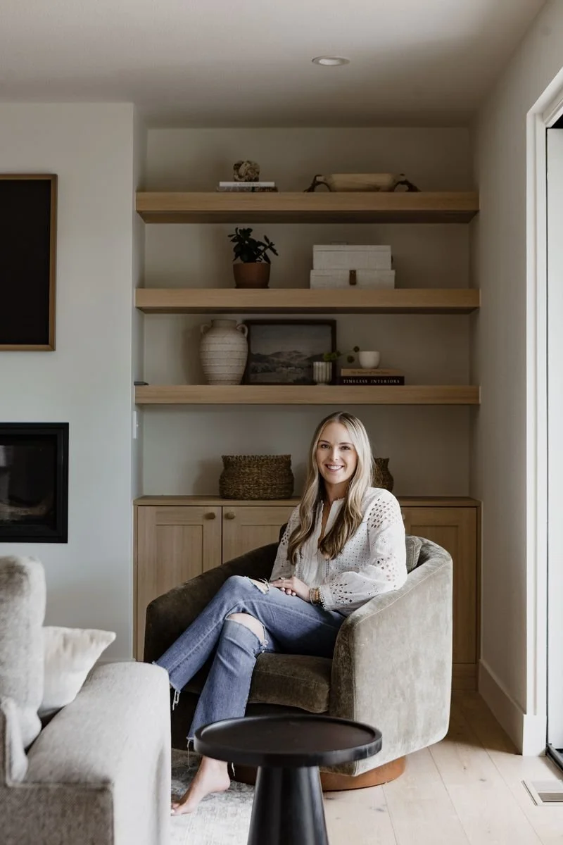 A woman with blonde hair and a white blouse sits on a beige armchair in a living room. She is smiling and has crossed legs, wearing ripped jeans. Behind her, there are wood shelves with decorative items like potted plants, books, and framed pictures. A cabinet is beneath the shelves, and a partial view of a fireplace is to the left.