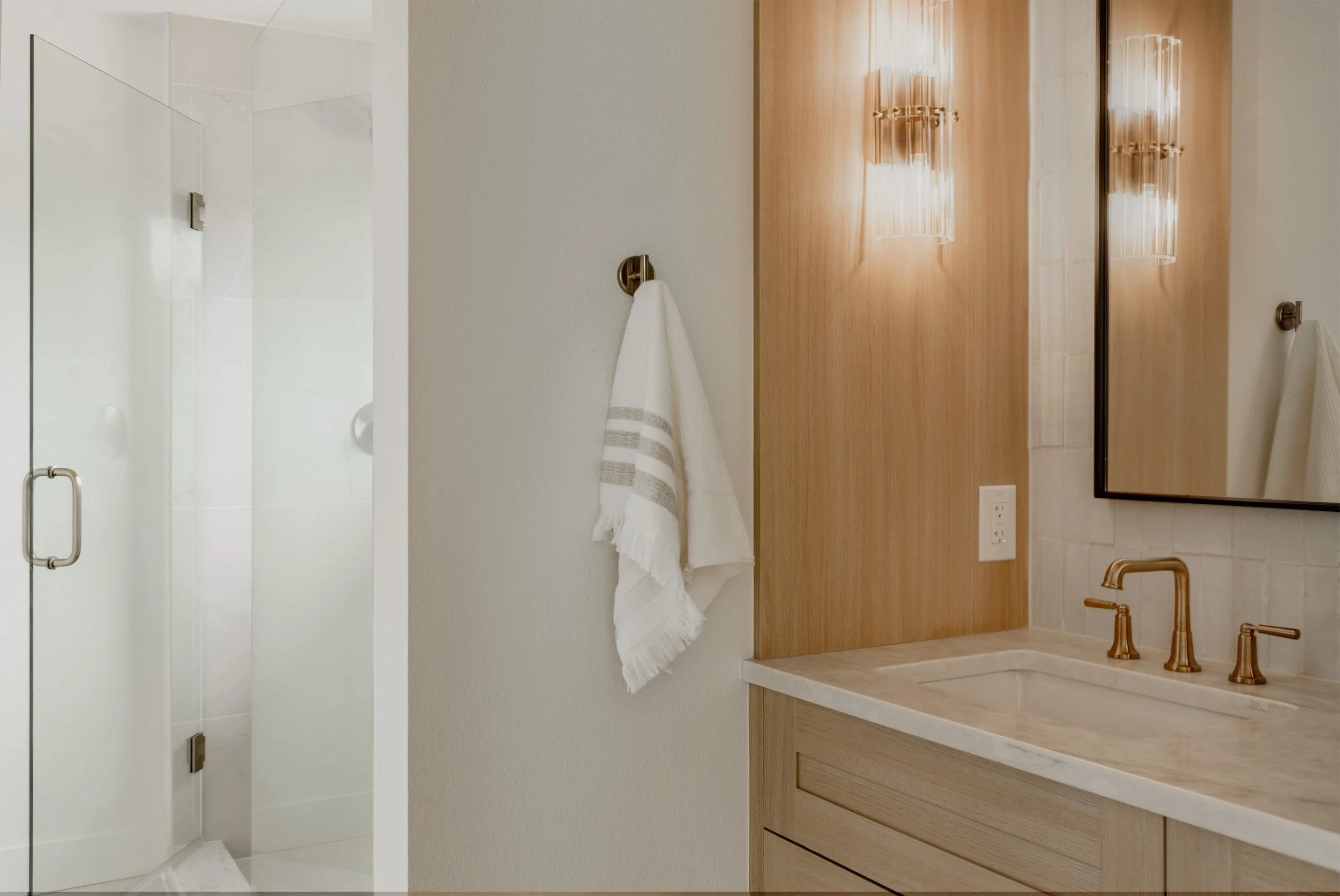 A bathroom with a walk-in shower enclosed with clear glass, a white towel hanging on a brass towel ring, a wooden vanity with a white marble countertop, brass fixtures, a large mirror, and wall-mounted lights.