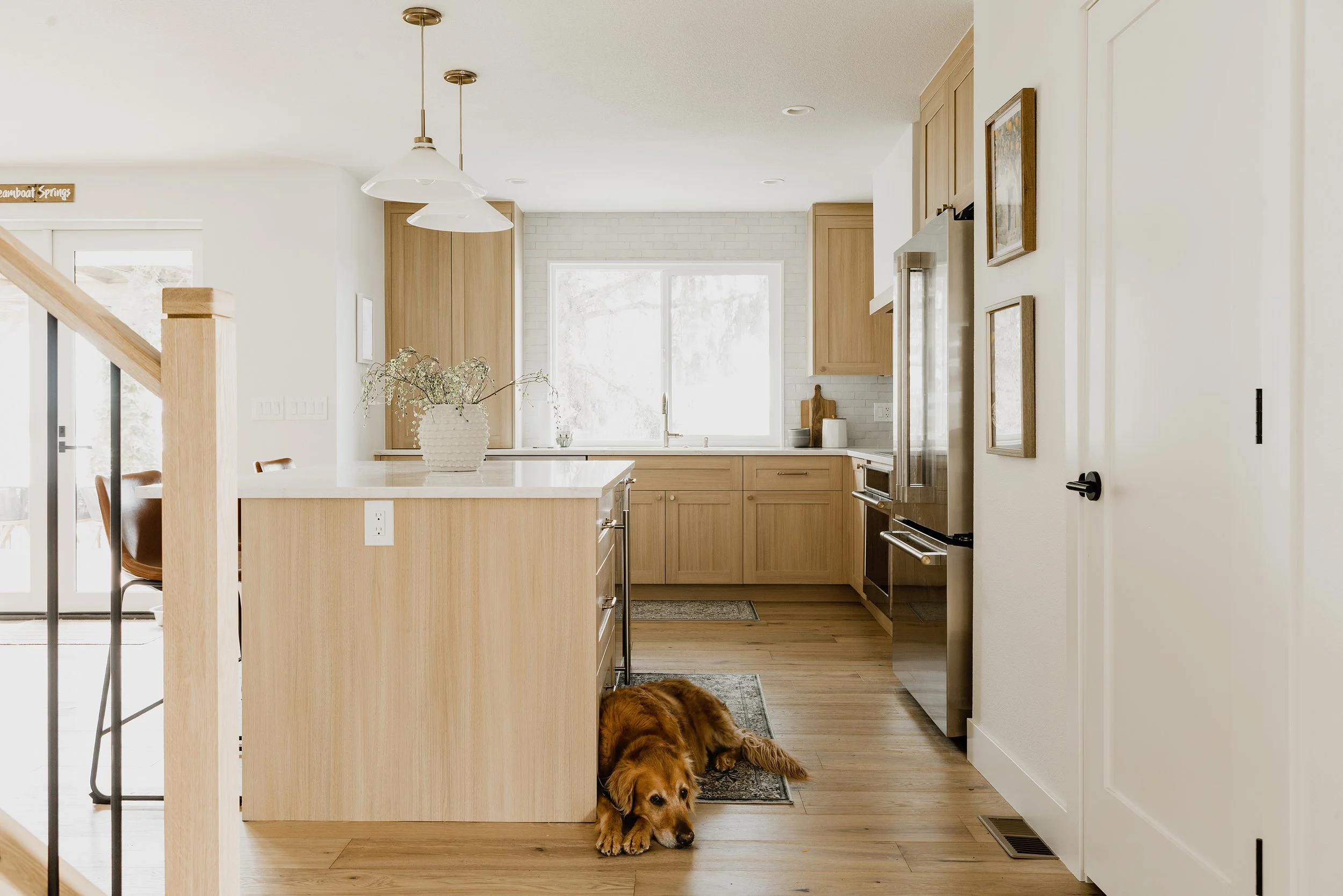 Open kitchen with light wood cabinets, white countertops, large window, hanging pendant lights, a decorative vase with flowers on the island, dog lying on the floor near the kitchen entrance.