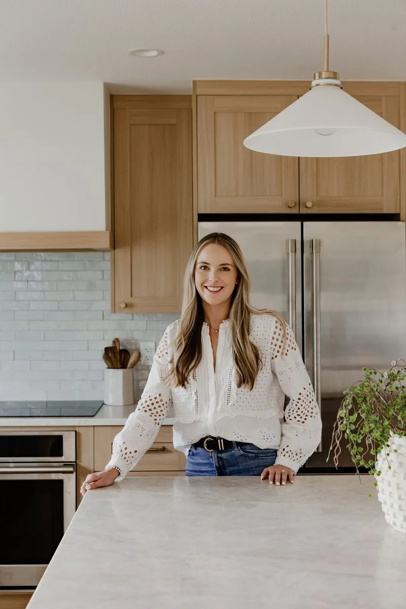 A woman with long blonde hair smiling in a modern kitchen, wearing a white eyelet blouse and jeans, standing behind a kitchen island with a potted plant on it.