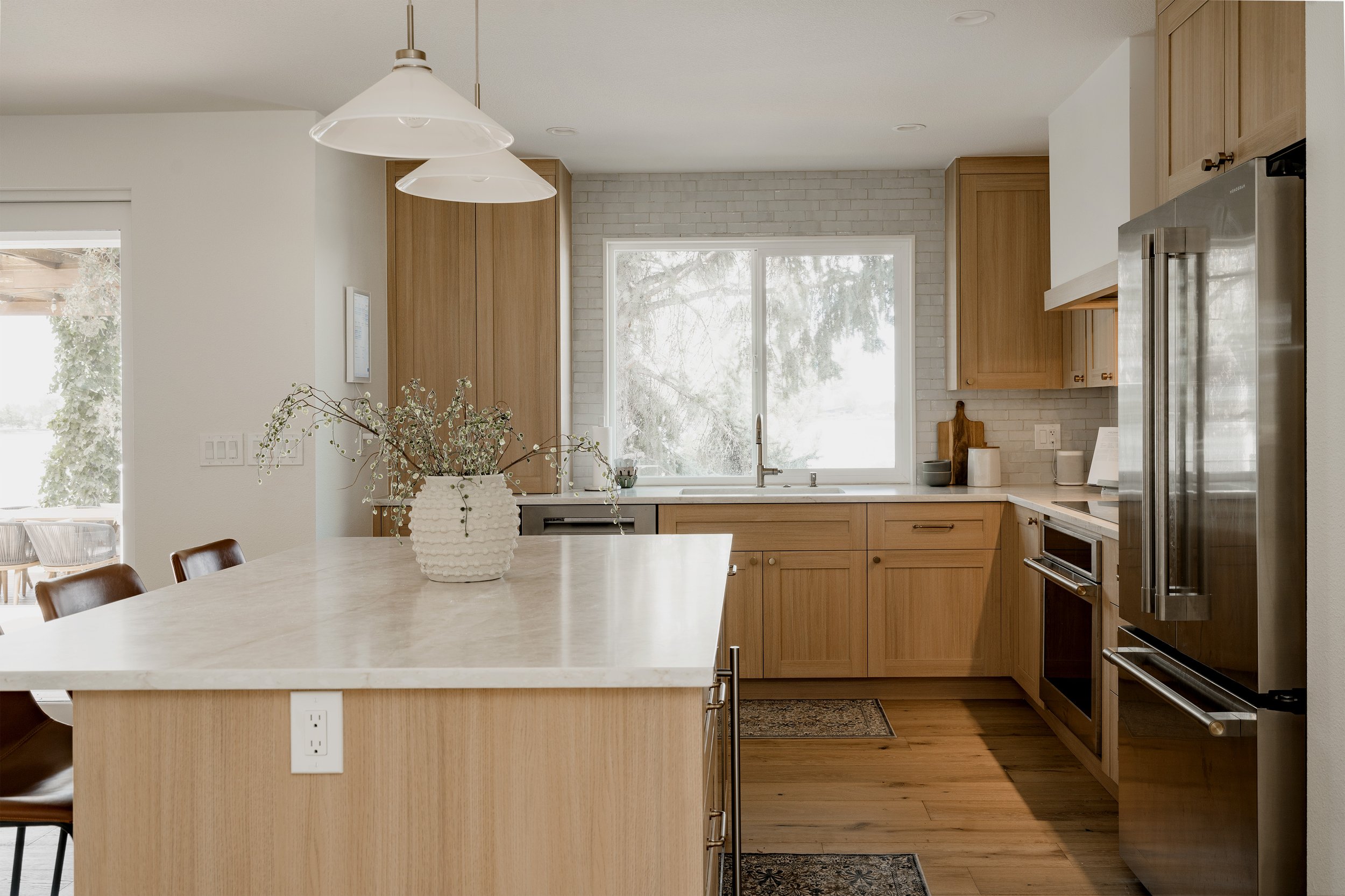 Modern kitchen with wood cabinets, white countertops, stainless steel appliances, large window, and a decorative white vase with branches on the island table.