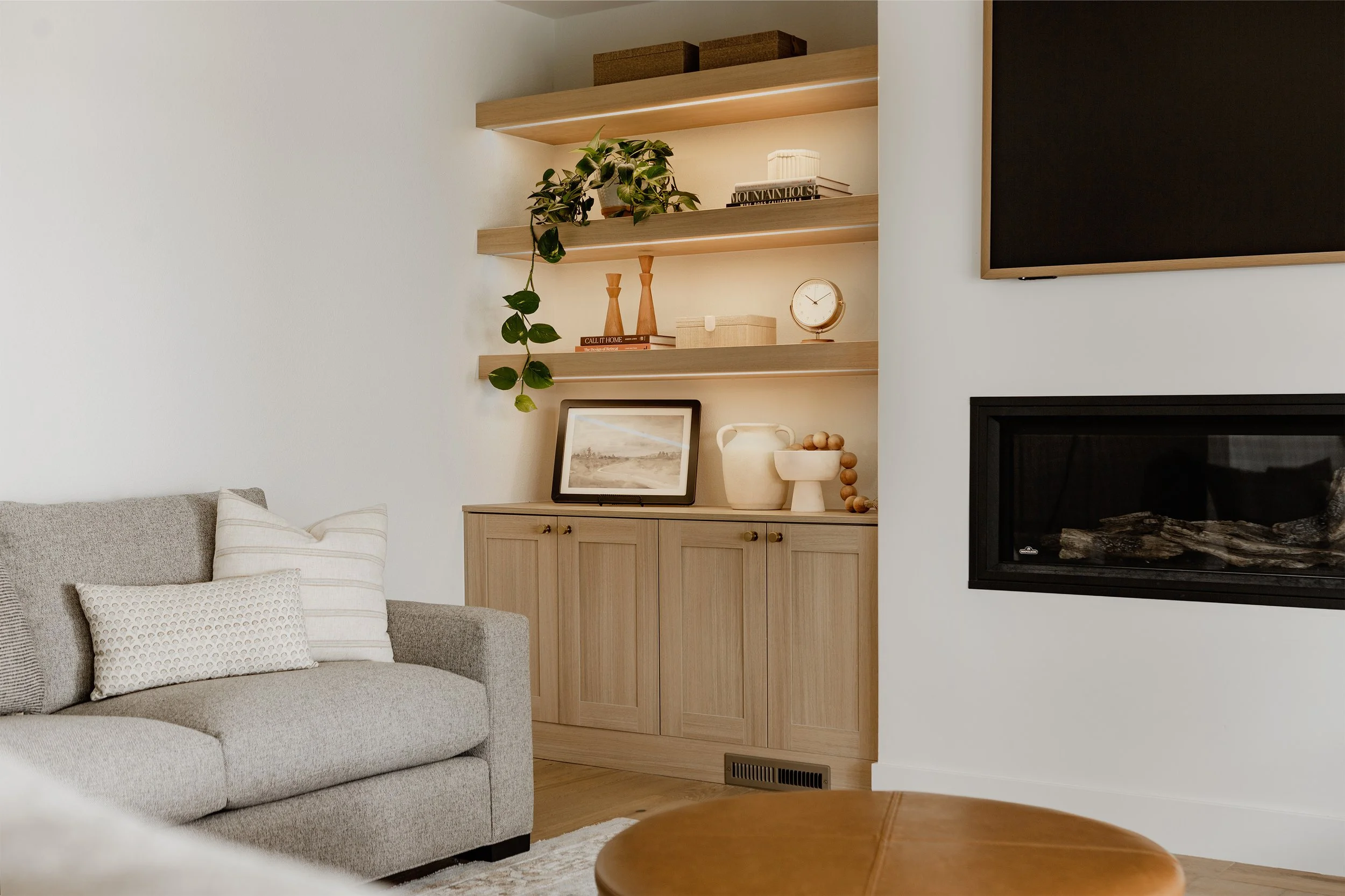 Living room with a beige sofa, decorative pillows, wall-mounted wooden shelves with plants and books, a framed picture, a vase, a clock, and a fireplace.