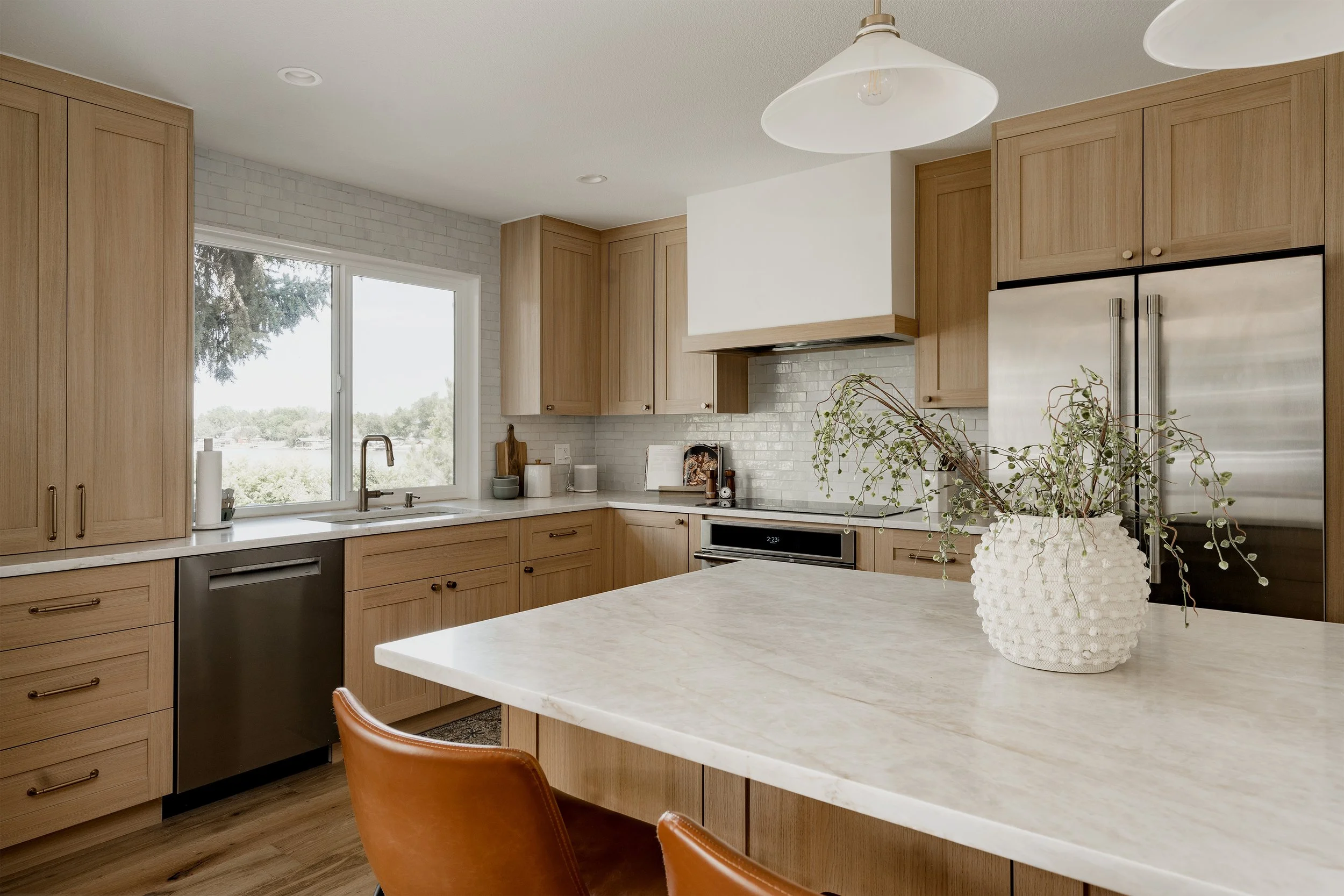 A modern kitchen with light wood cabinets, a white countertop, a stainless steel refrigerator, and a window overlooking trees outside. There is a potted plant on the counter, and ceiling lights.