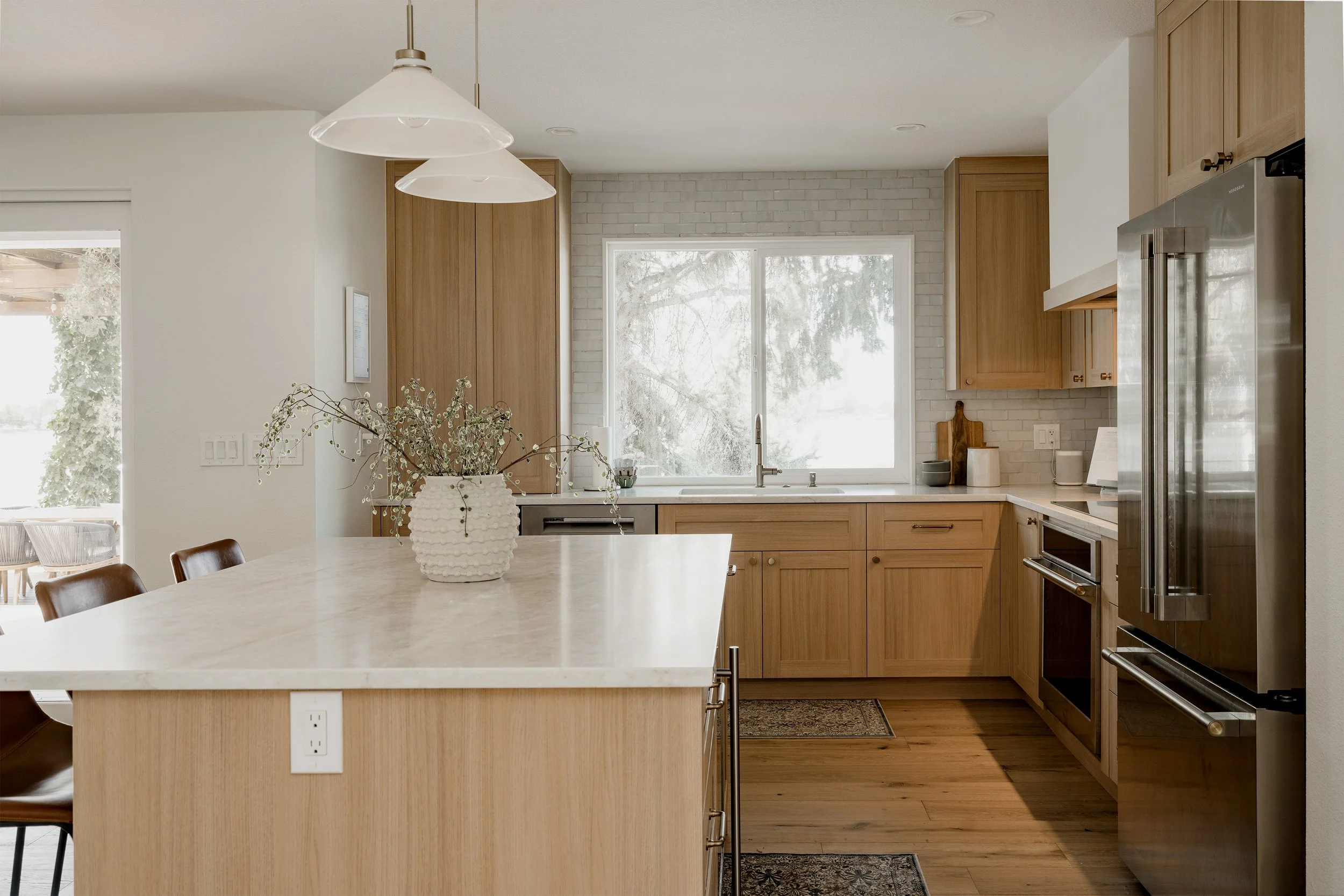 Modern kitchen with wooden cabinets, stainless steel appliances, large window, and a white countertop island with a decorative vase of dried flowers.
